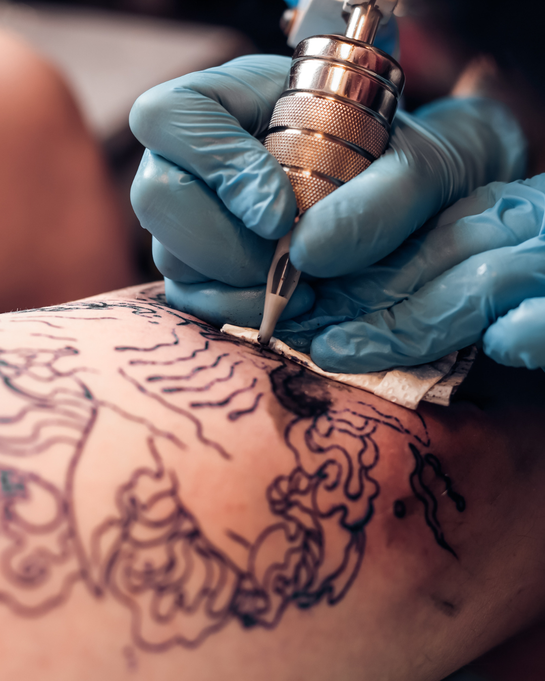 A tattoo artist in blue gloves using a tattoo machine to ink a detailed tribal design on a person's arm.