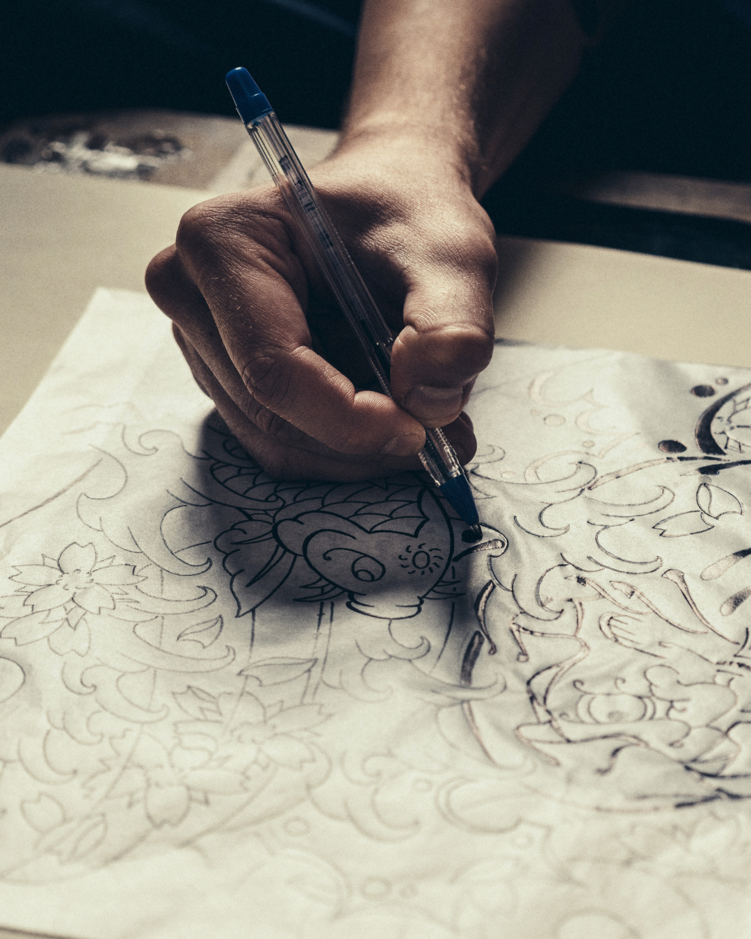 A hand holding a pen and drawing intricate floral and animal designs on a piece of paper.