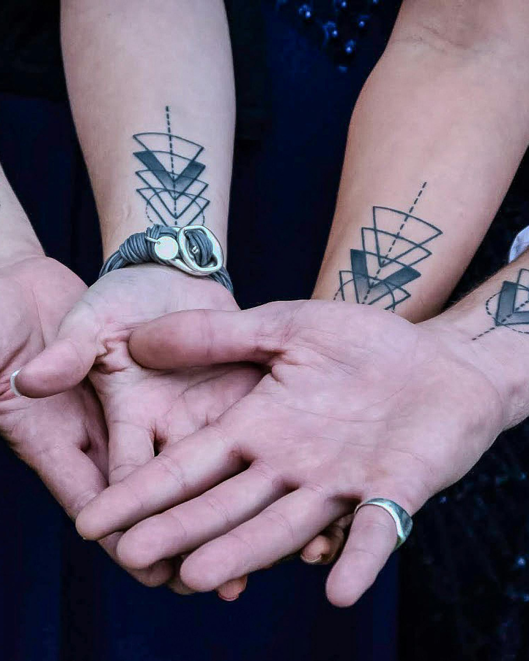 Close-up of two people holding hands, with four geometric tattoo designs on their forearms showing overlapping shapes and a dotted line, and wearing rings and a bracelet.