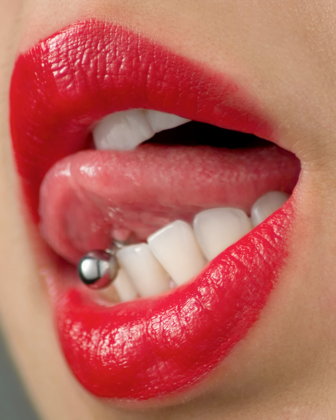 Close-up of a person's lips with bright red lipstick, tongue slightly sticking out, and white teeth visible, with a tongue piercing.