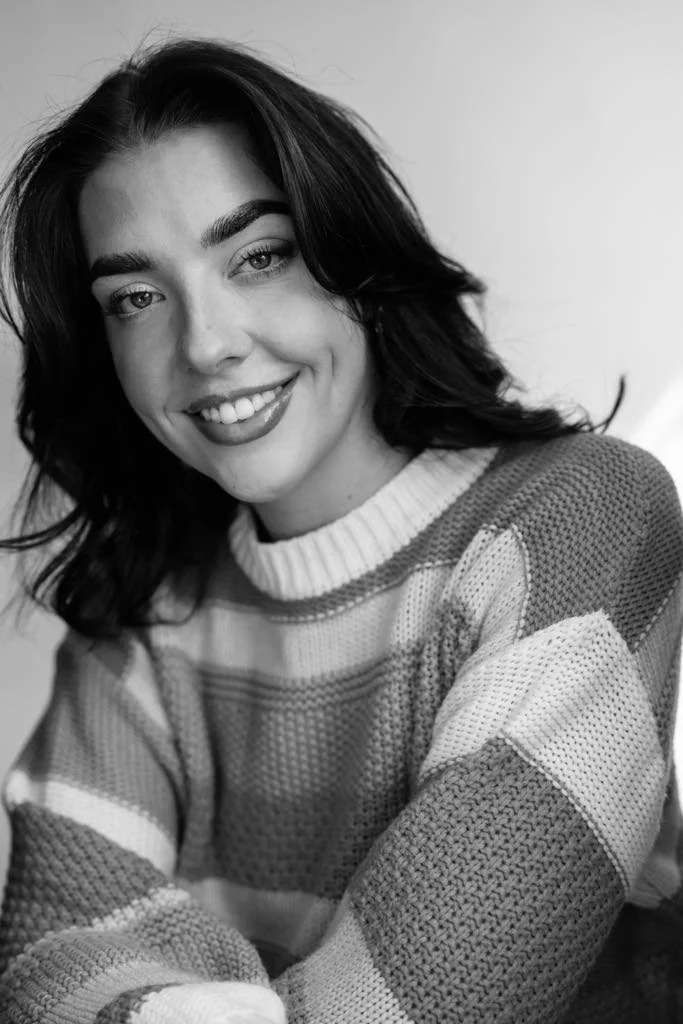 Black and white photo of a young woman smiling, with shoulder-length dark hair, wearing a striped knitted sweater.
