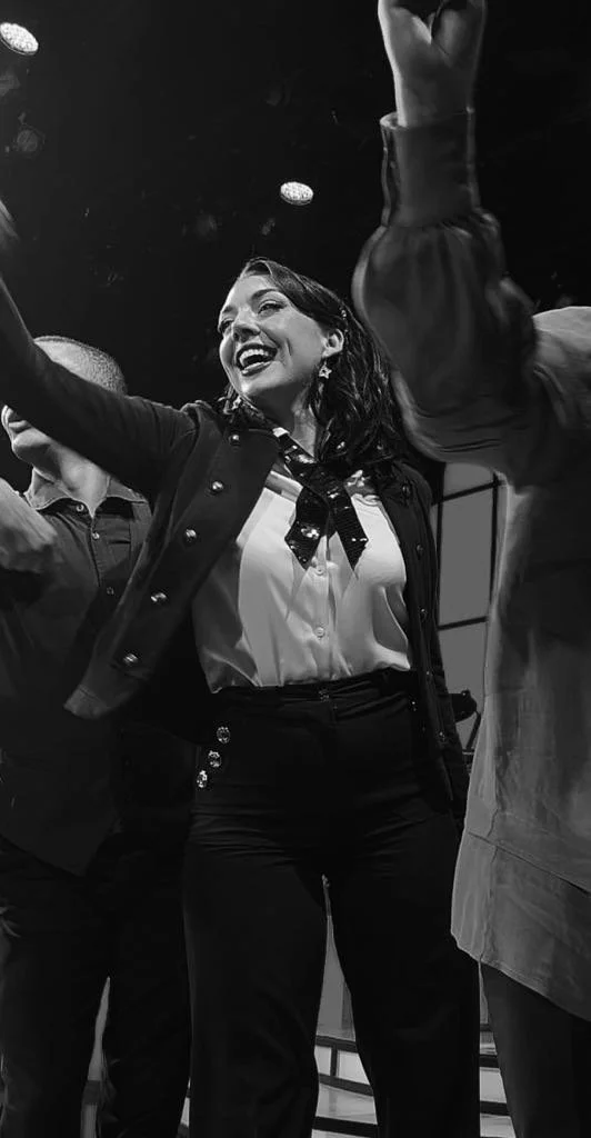 A woman smiling and dancing in a group, wearing a blazer, blouse, and necktie, with arms raised in celebration.