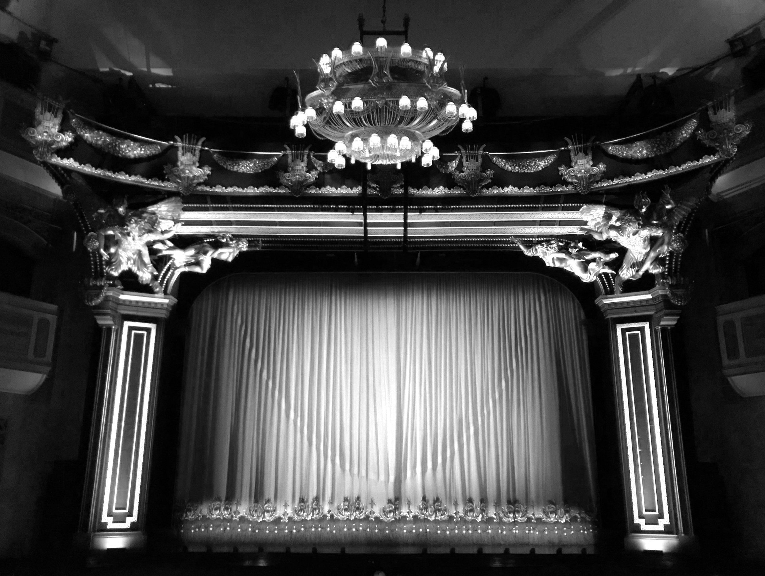 Black and white photo of a theater stage with a closed curtain, ornate columns, and decorative sculptures and chandeliers above.