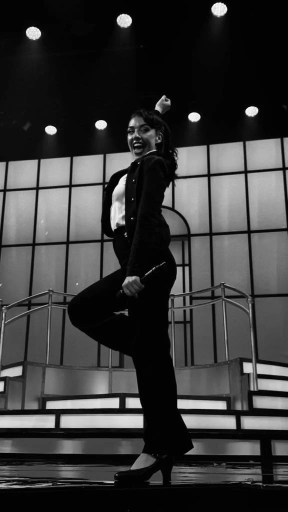 A woman on stage, smiling and striking a playful pose, holding a microphone in one hand, with a backdrop and stage lights overhead.