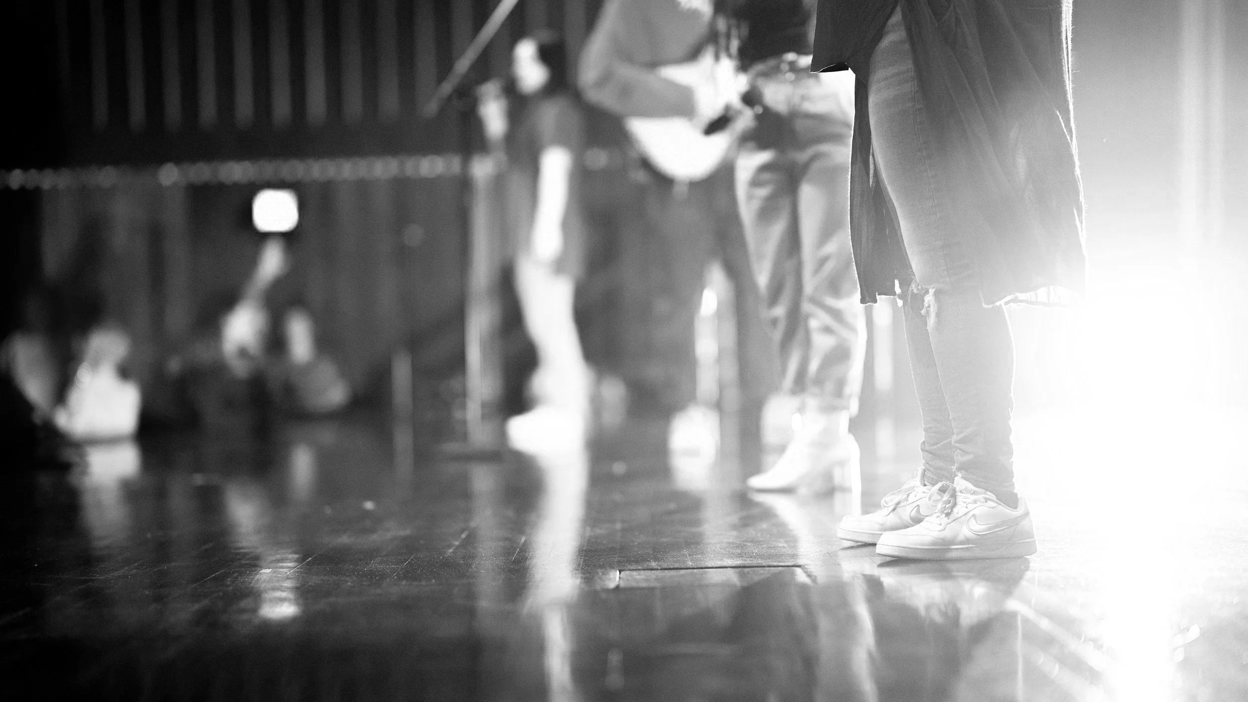 People standing in a row, viewed from the knees down, in a room with wooden floor, some wearing sneakers and some wearing high heels. Bright light in the background.