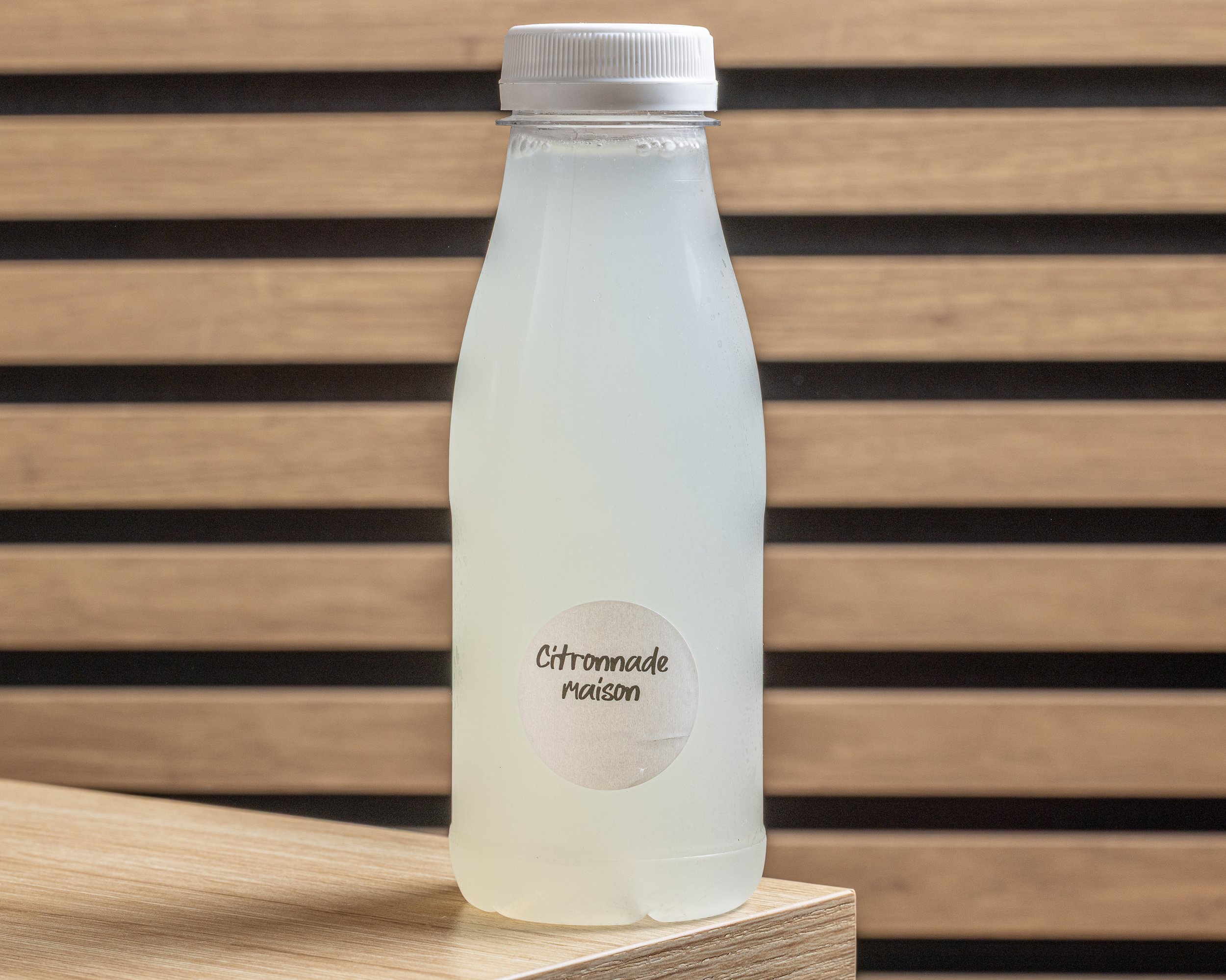 Une bouteille de citronnade maison avec un bouchon blanc, posée sur une surface en bois, avec un fond en lamelles de bois horizontal.