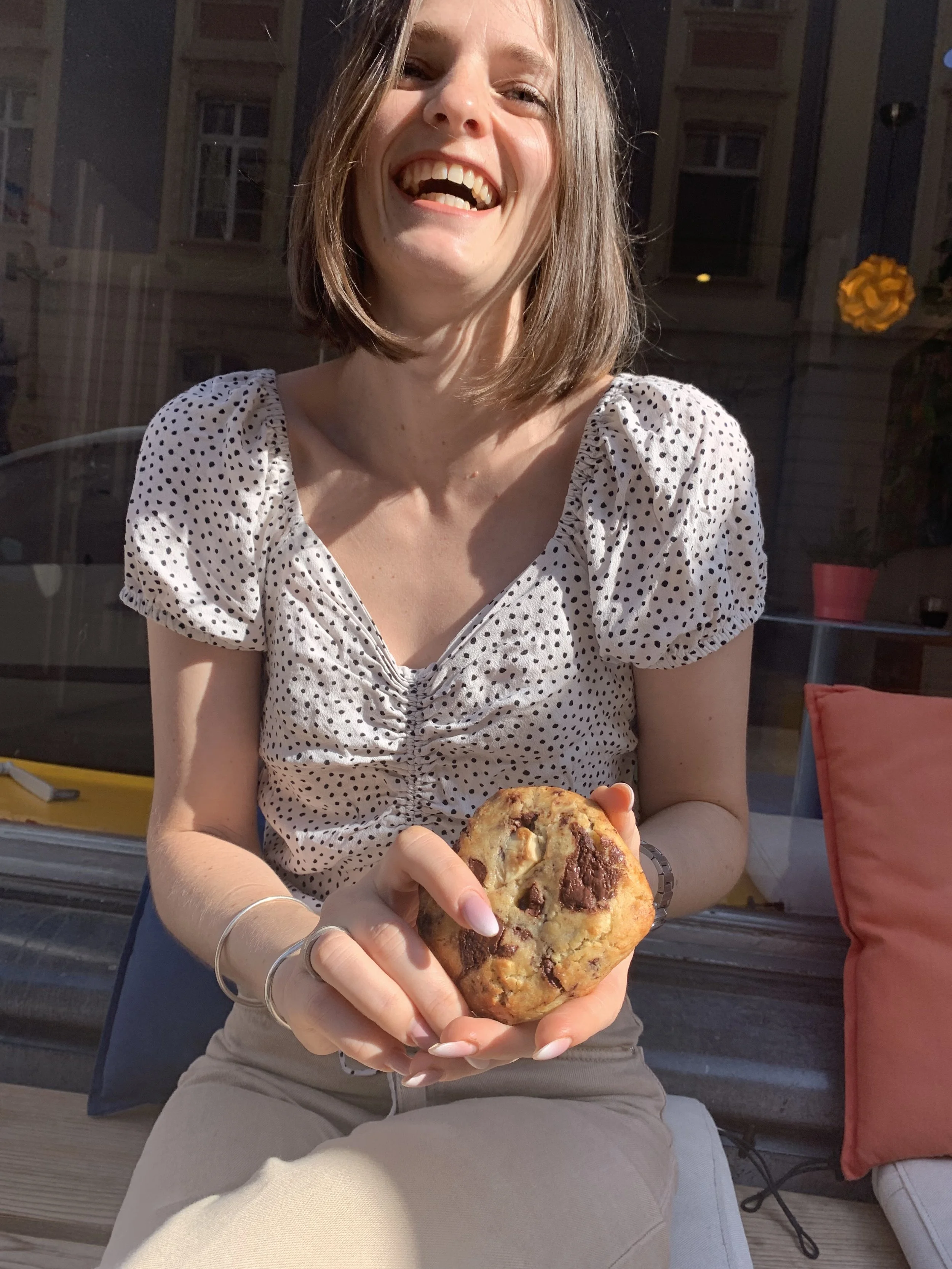 Une femme souriante, assise à une table, tient un gros cookie aux pépites de chocolat, en plein jour.