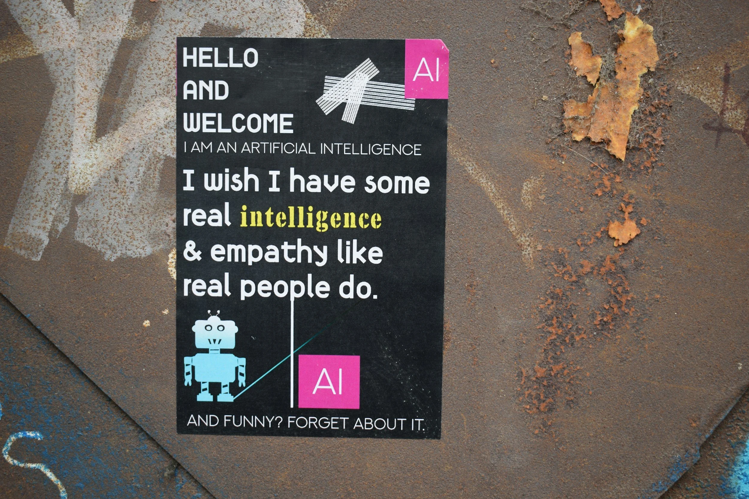A black poster with white, yellow, and pink text features a robot illustration and a white abstract design. The poster reads 'HELLO AND WELCOME I AM AN ARTIFICIAL INTELLIGENCE I wish I have some real intelligence & empathy like real people do. AND FUNNY? FORGET ABOUT IT.'