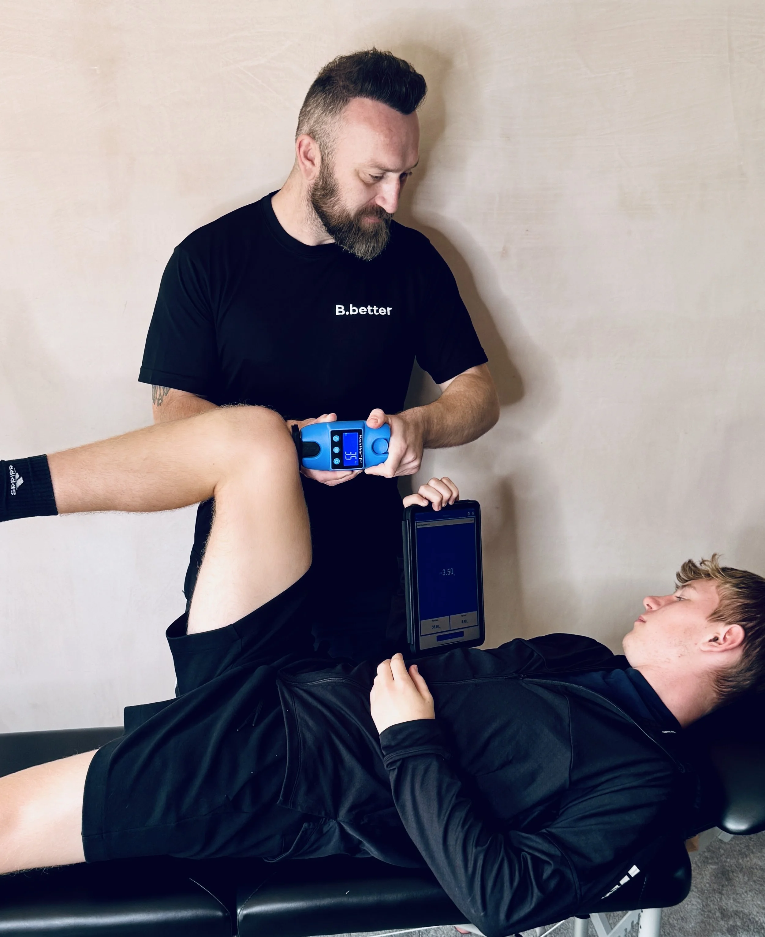 A physical therapist performs a leg flexibility test on a male patient lying on a treatment table, using a handheld device and a tablet for assessment.
