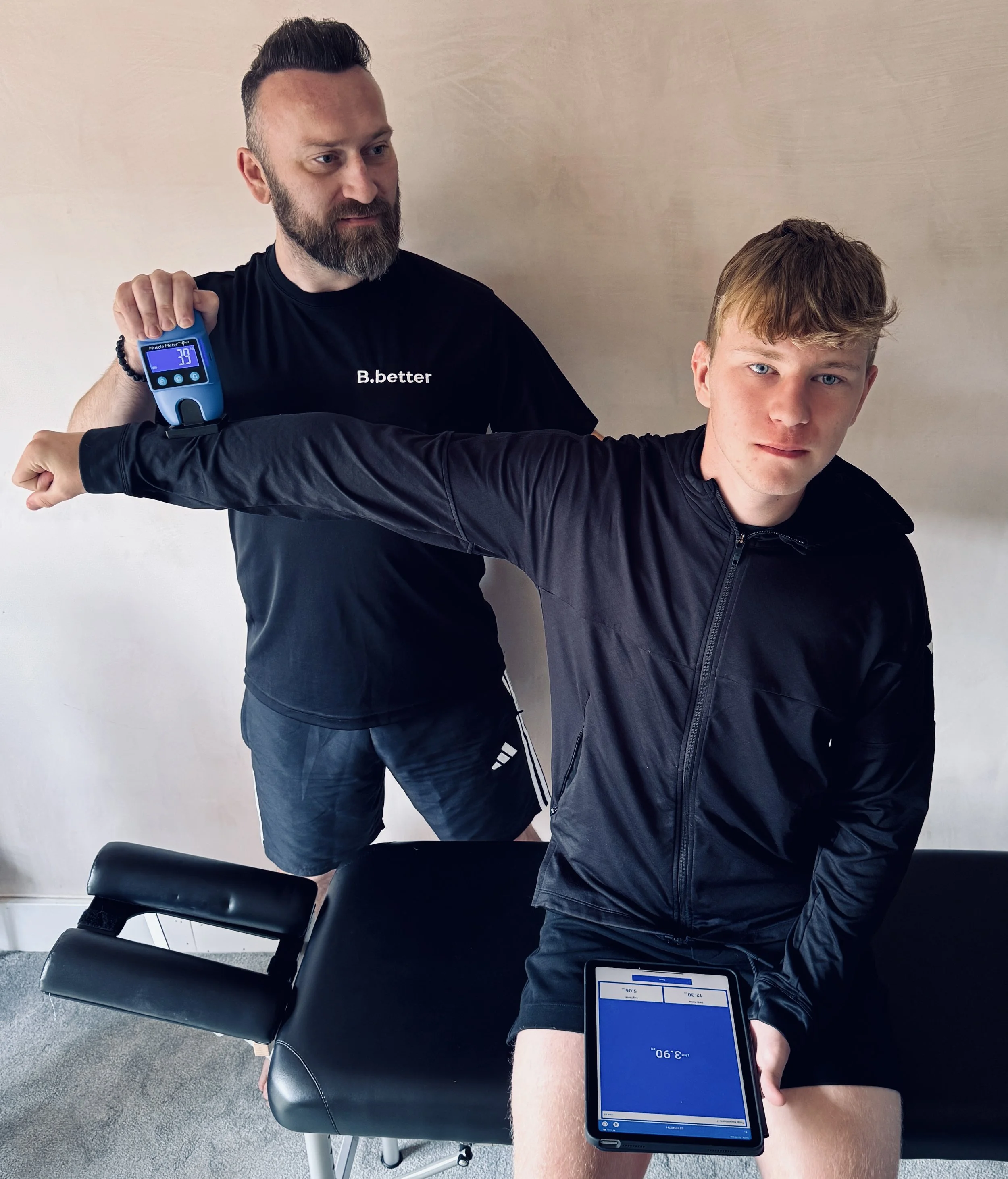 A man with a beard conducting a muscle test on a young man with blond hair and a black jacket using a muscle meter and tablet on a massage table.