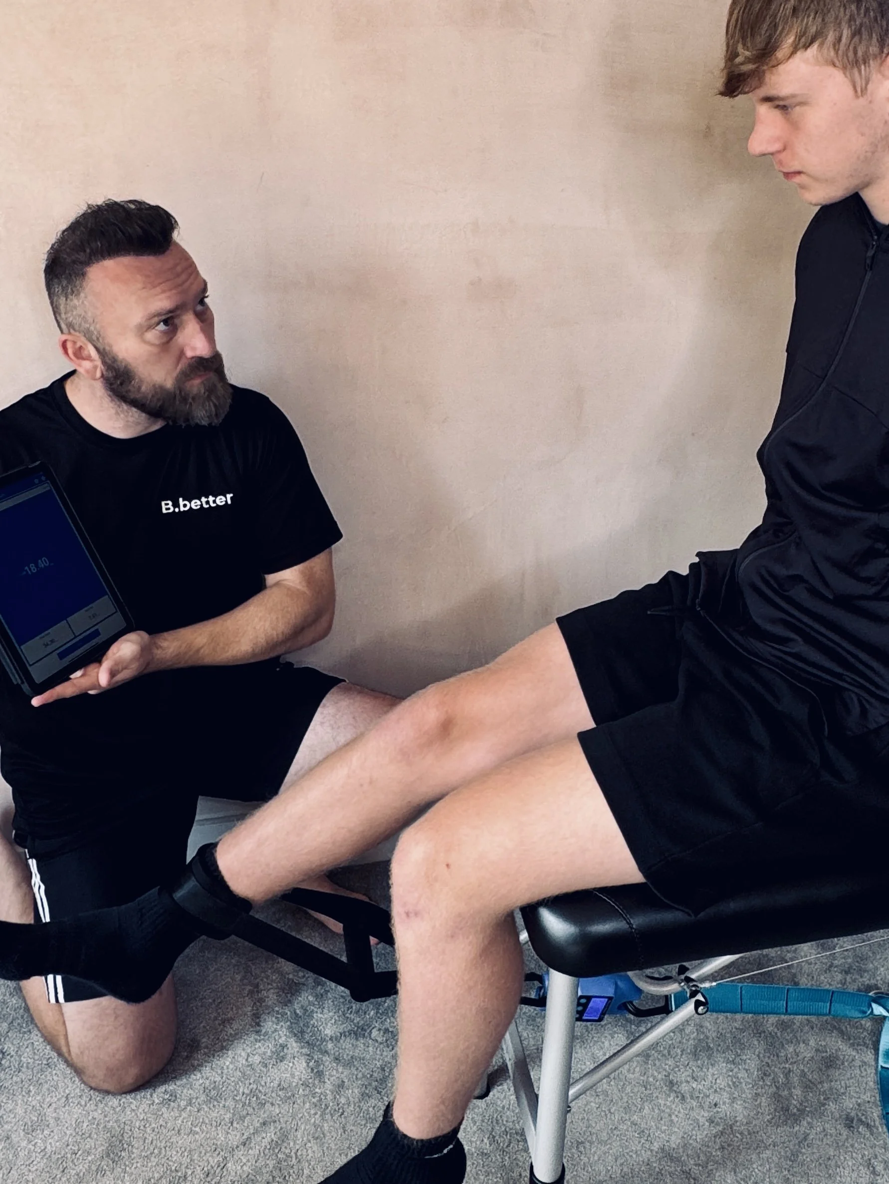 A man sitting on a black chair with his right leg extended, while another man kneels in front of him, holding his ankle. The man on the chair appears to be undergoing a physical examination or therapy. The kneeling man is wearing a black shirt, and the man on the chair is wearing a black long-sleeve shirt and shorts.