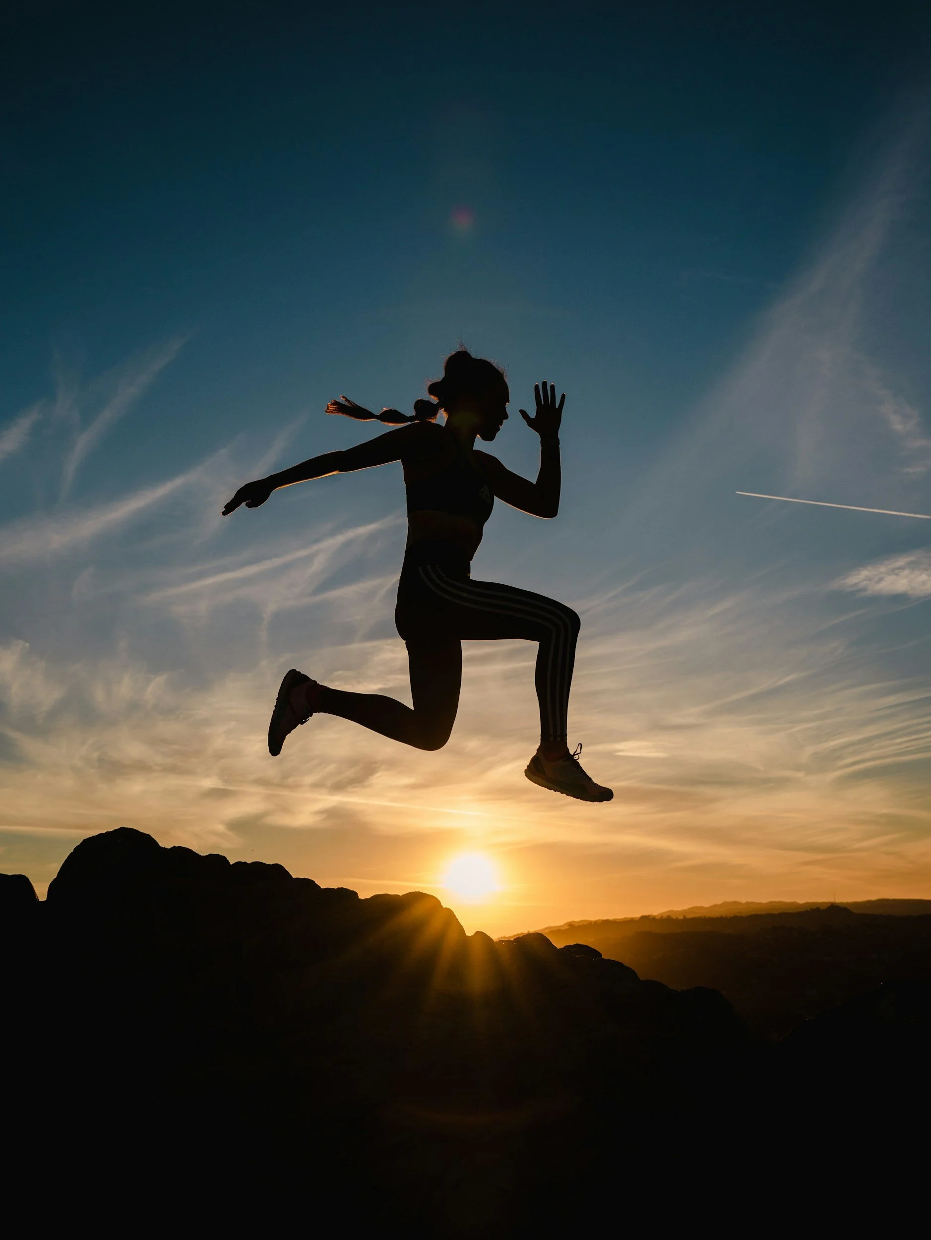 Silhouette of a woman running on rocky terrain at sunset with a clear sky and contrails.