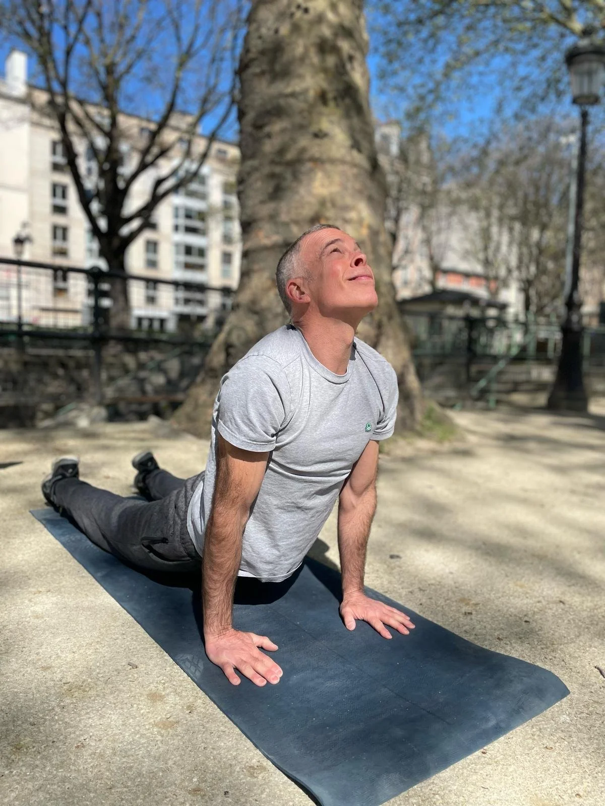 Man practicing yoga outdoors on a mat, in a yoga pose called upward dog, with a large tree and urban buildings in the background.