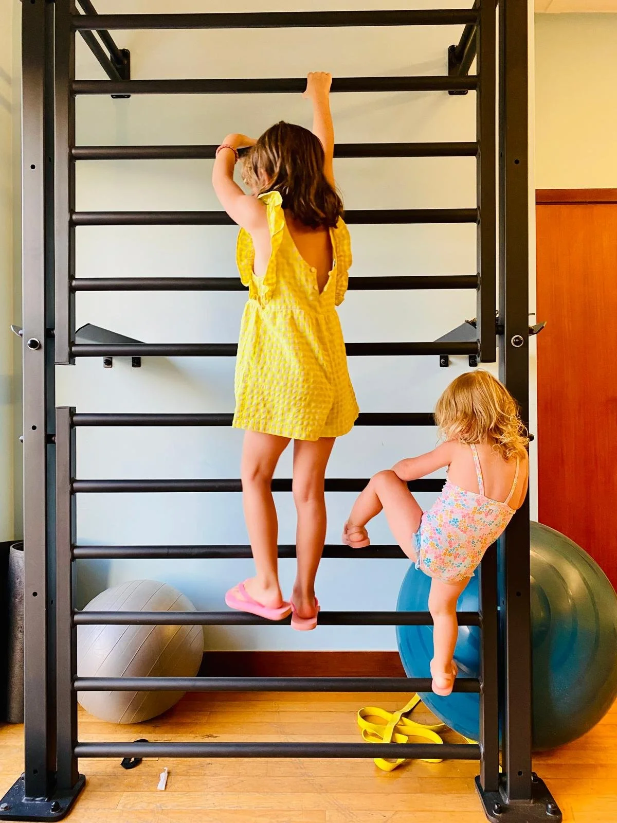 Two young girls climbing on a gym climbing frame in a play area, with exercise balls and foam rollers on the floor.