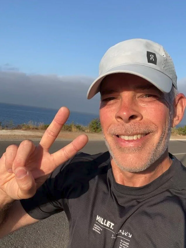 Smiling man in athletic wear taking a selfie outdoors near water, making a peace sign with his fingers, wearing a light-colored cap and black shirt, with a road and ocean in the background.