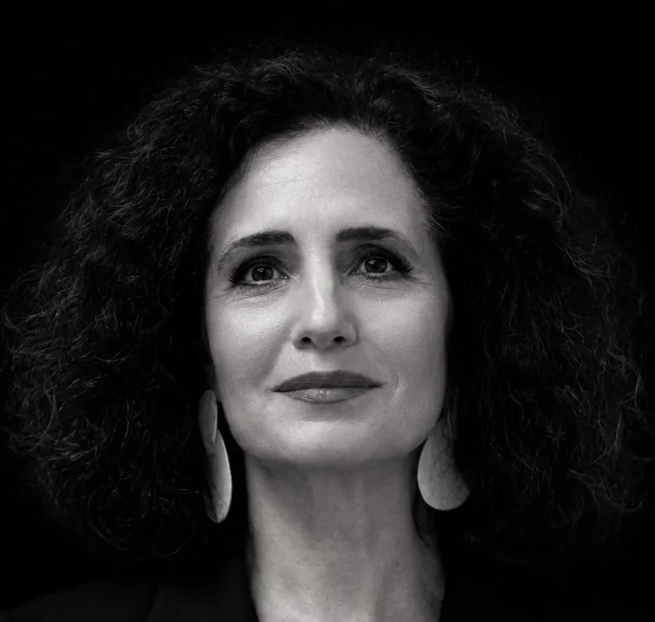 Retrato de uma mulher com cabelo cacheado e escuro, usando blusa de blazer com listras finas, brincos grandes, olhando para cima, em preto e branco.
