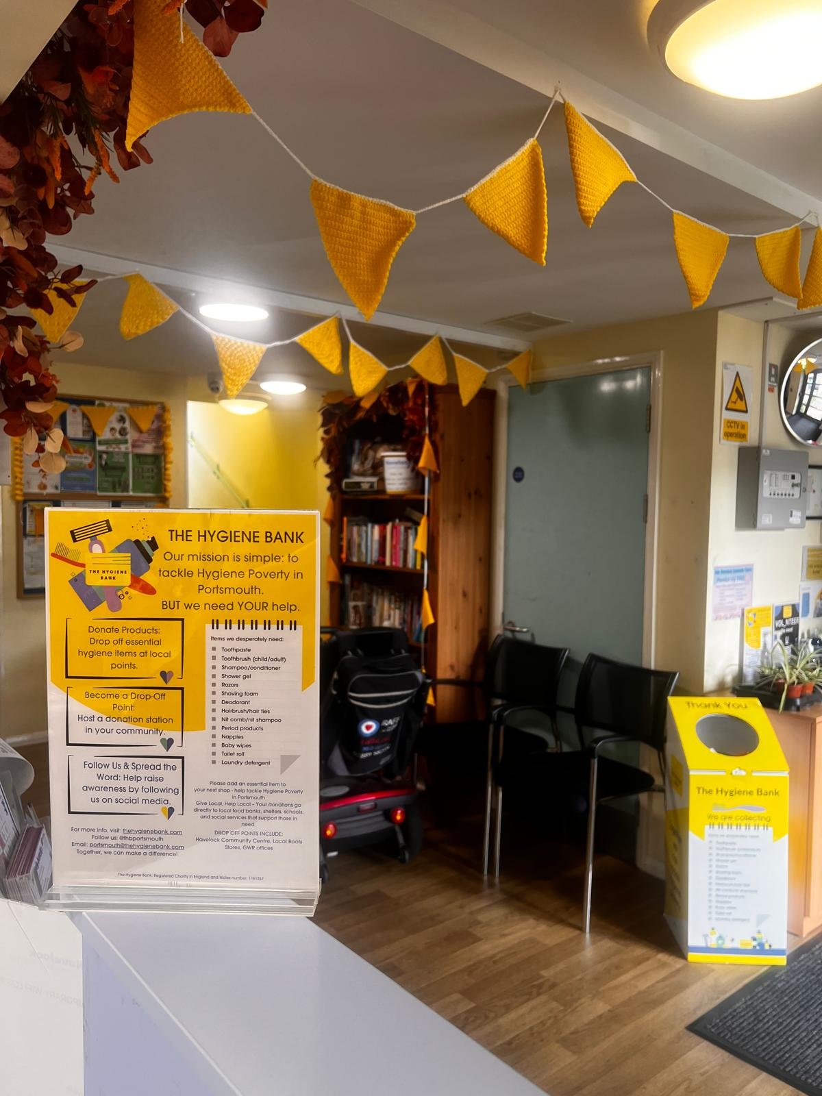 Interior of a community center with yellow hygiene bank donation bin, yellow triangular bunting, chairs, wooden shelves, and information posters.