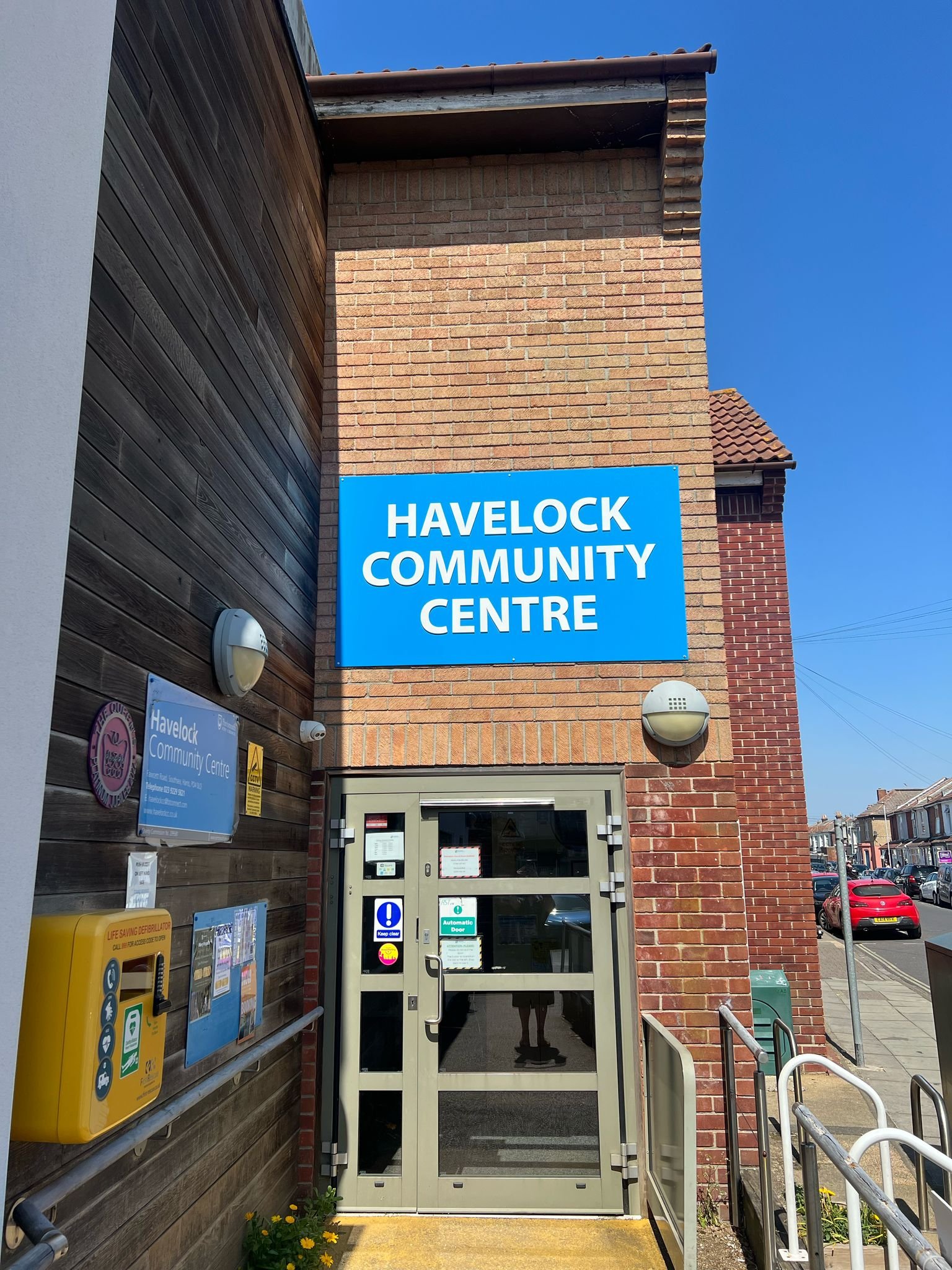 The entrance to Havelock Community Centre with a blue sign displaying its name, a glass door, and surrounding brick and wooden exterior walls under a clear blue sky.