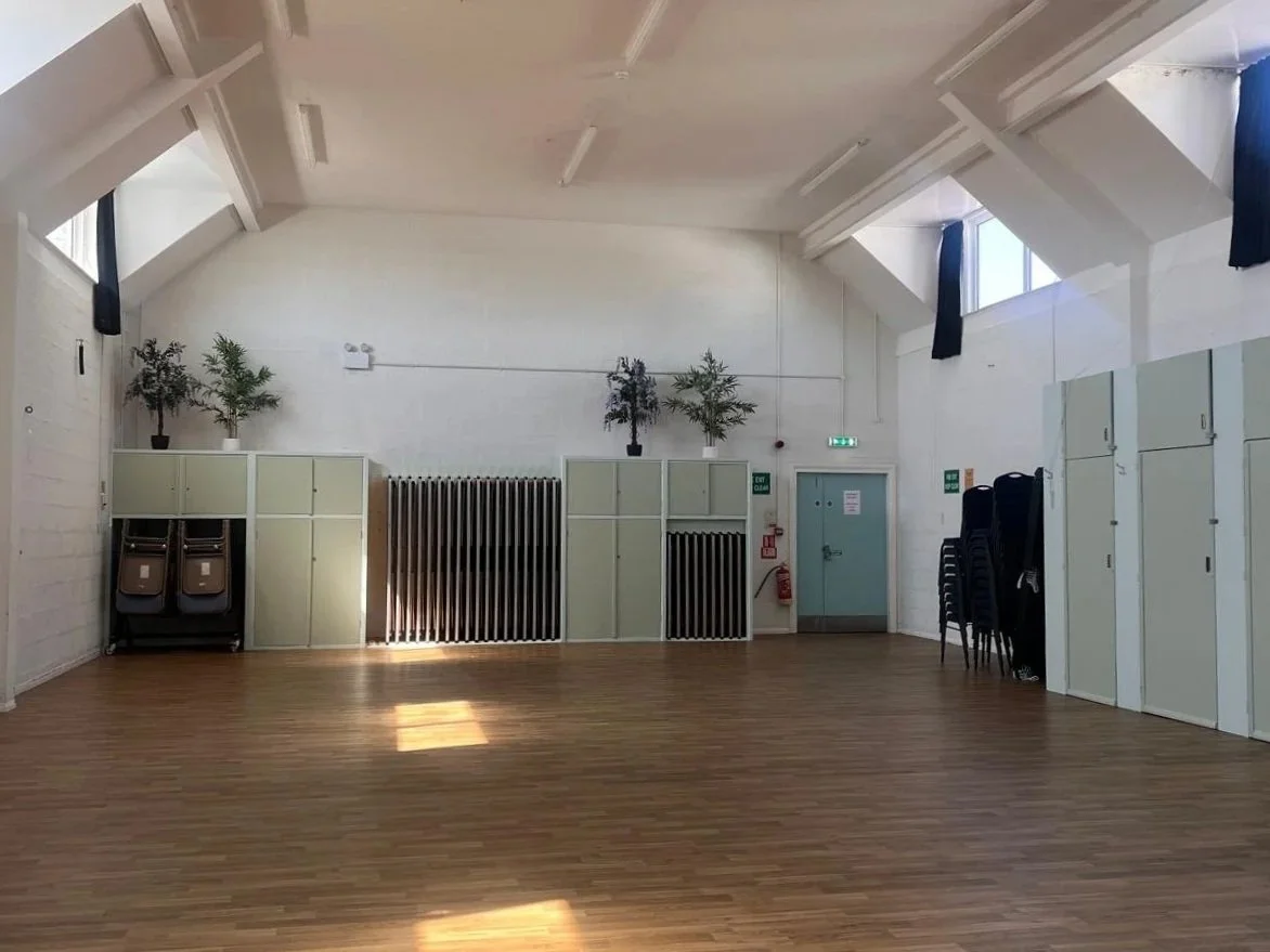 An empty multipurpose room with wooden floor, white walls, and high ceiling with skylights. There are two potted plants on cabinets, chairs stacked against the wall, and a closed door with signs and fire extinguisher nearby.