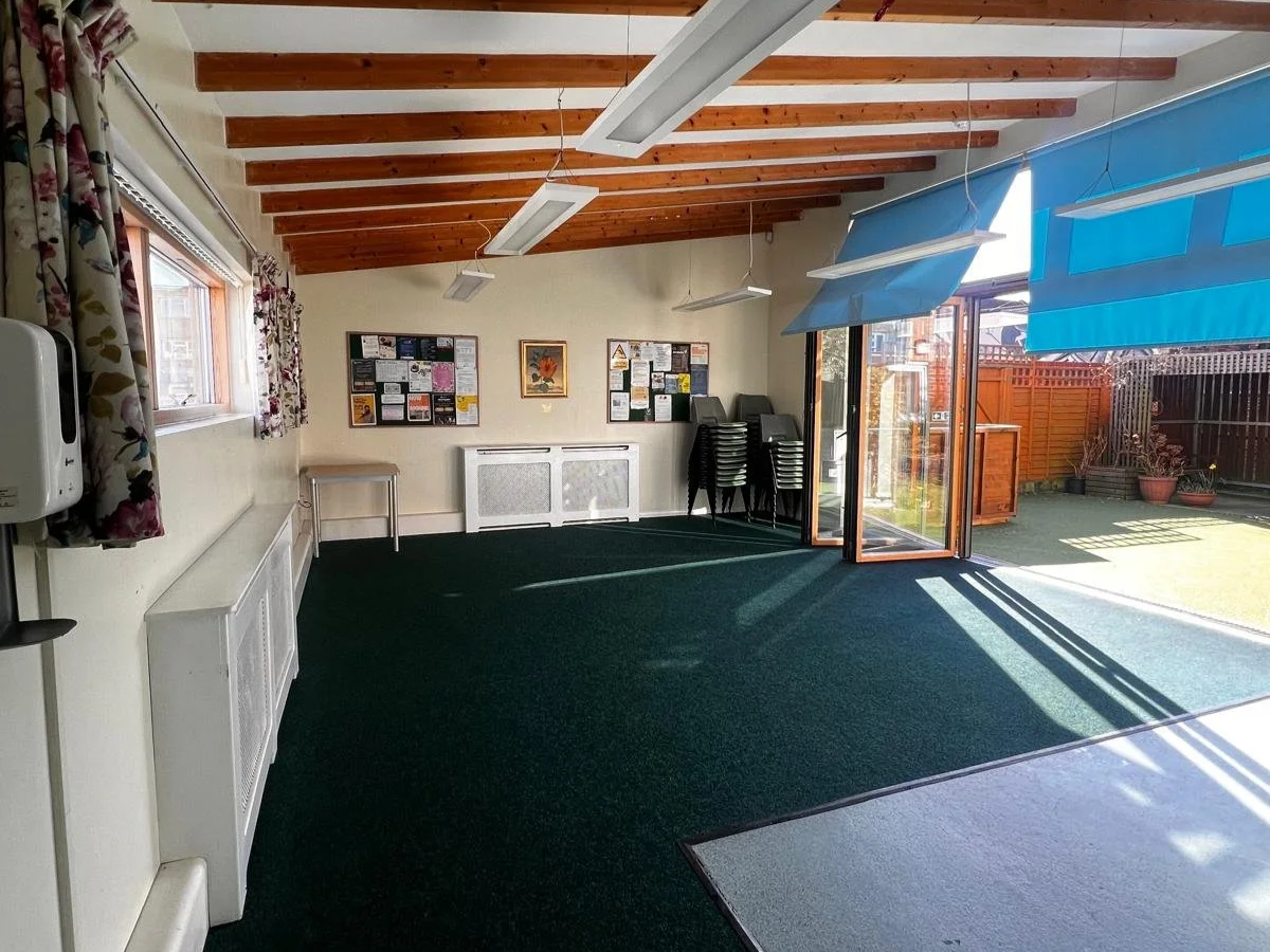 Sunlit indoor space with green carpet, stacked chairs, and open sliding glass door leading to a fenced backyard with potted plants.