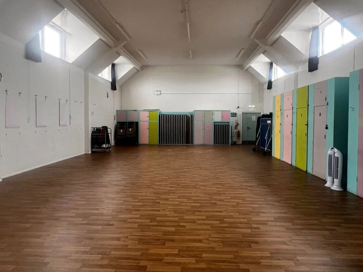 Empty room with wooden floors, lockers on the right wall painted in pastel colors, and folding chairs stacked on the left side, with high windows providing natural light.