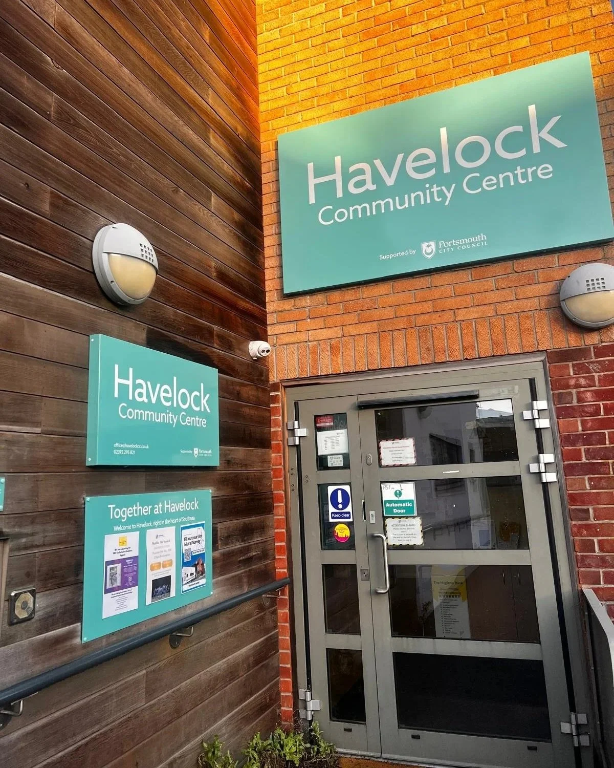 Exterior view of Havelock Community Centre showing a large teal sign and smaller teal sign on the wall, a glass door with various stickers, and two wall-mounted outdoor lights, with a brick and wooden wall background.