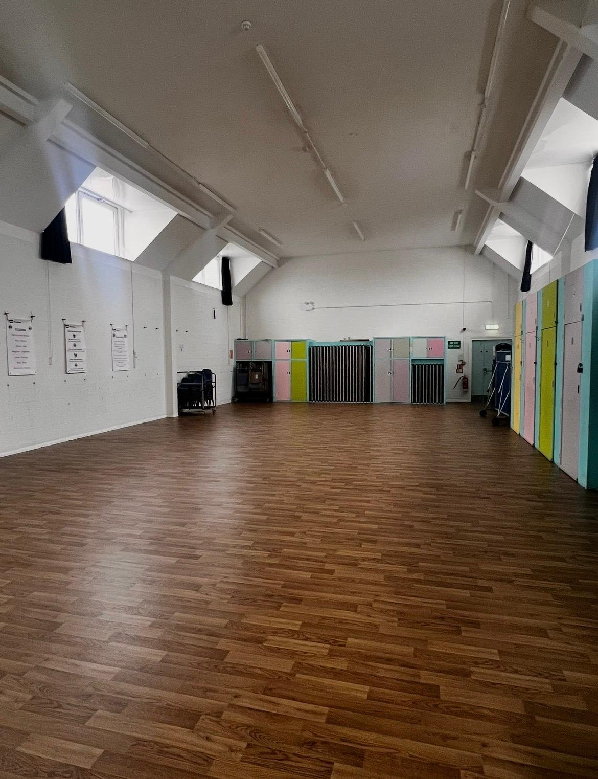Empty room with wooden flooring, white walls, windows high on the sloped ceiling, lockers on the back wall, and some chairs and cleaning supplies on the right side.