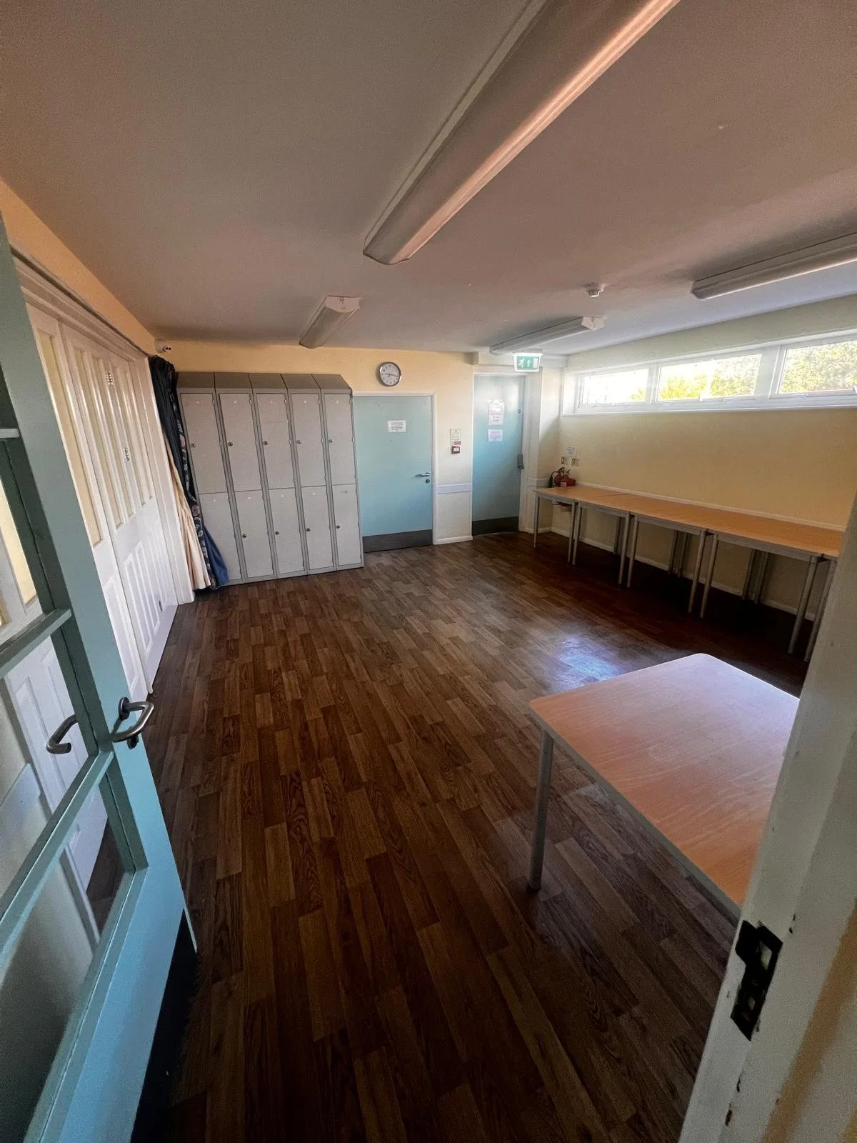Empty classroom with wooden floor, grey lockers, a door, and large window letting in natural light.