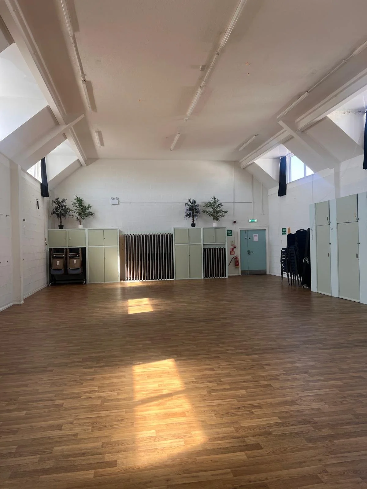 Empty room with wooden flooring, pastel-colored lockers on the right, and some black carts and a fan on the left side.