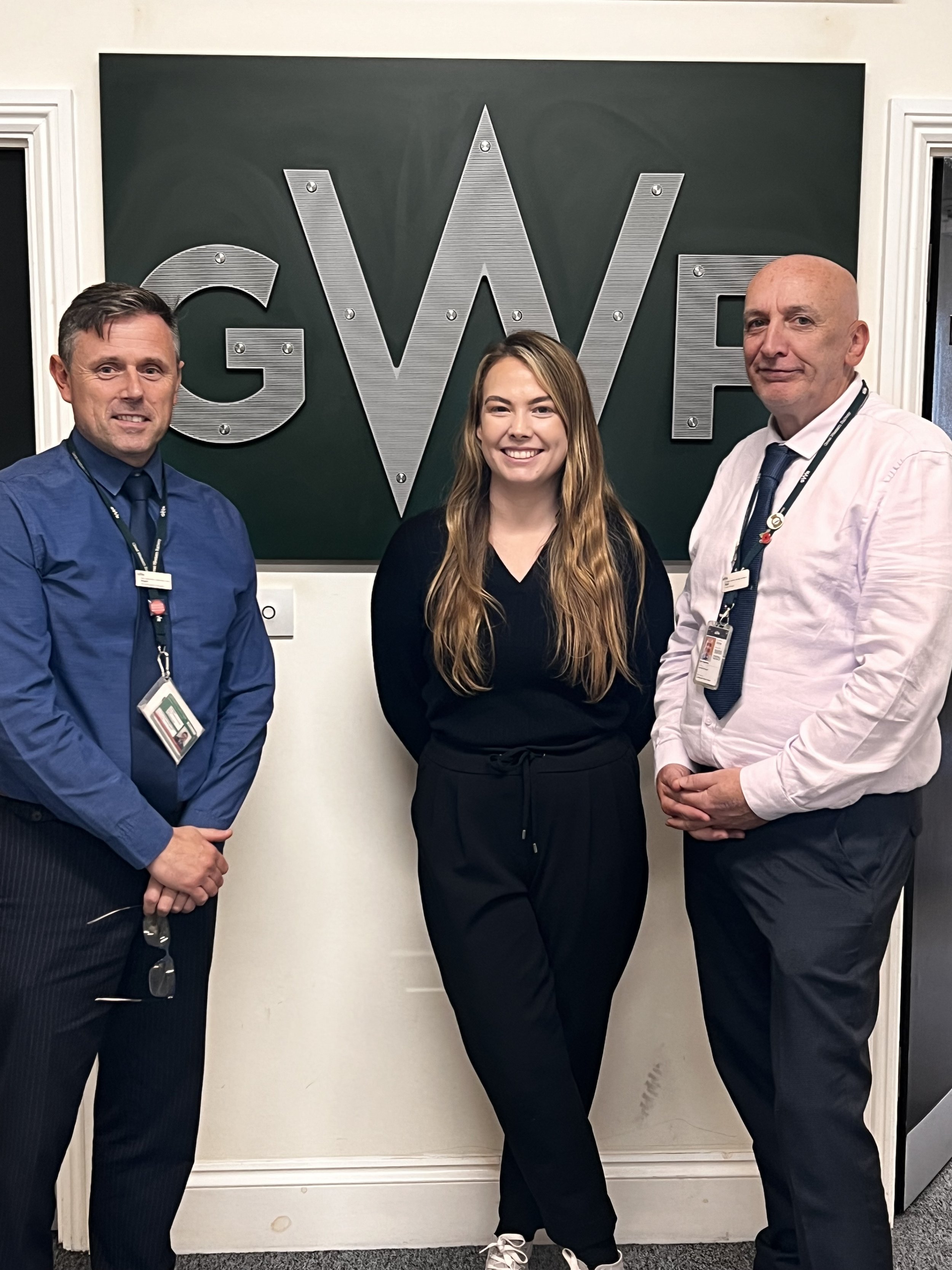 Three people standing in front of a wall with large metallic letters that read 'GWE.'