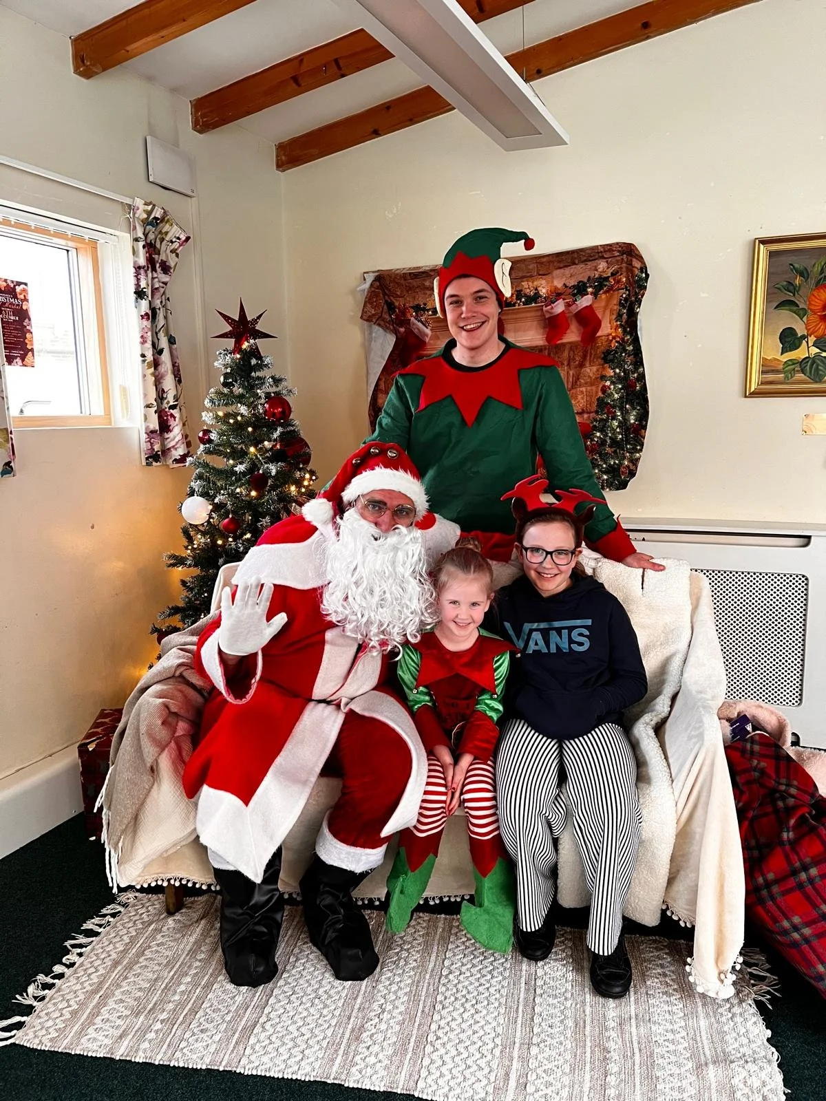 A group of four people, dressed as Santa Claus and elves, posing with two children on a holiday-themed sofa in a decorated room with a Christmas tree, stockings, and festive decorations.