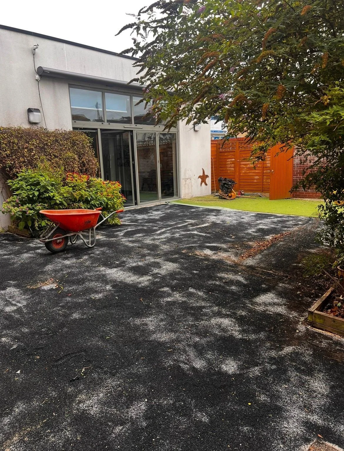 Our Garden having a makeover thanks to National Lottery funding- new astro turf in coming a red wheelbarrow, green bushes, a tree, a wooden fence, and a building with large glass sliding doors.