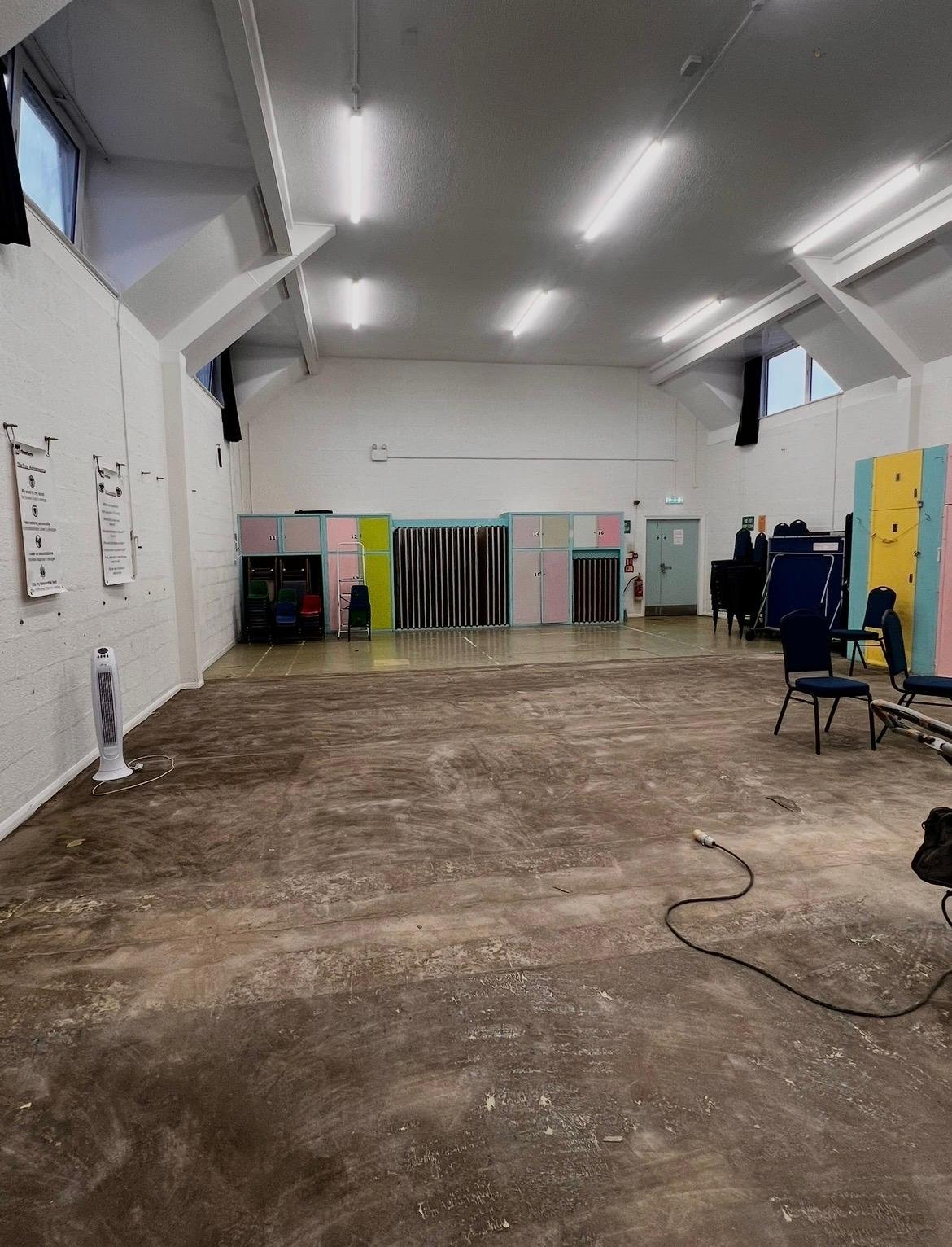 Work underway in The Bennett Hall at Havelock with Commercial Building Solutions installing new flooring with brown flooring, white walls, colorful lockers at the back, chairs on the right, a fan, and some equipment.