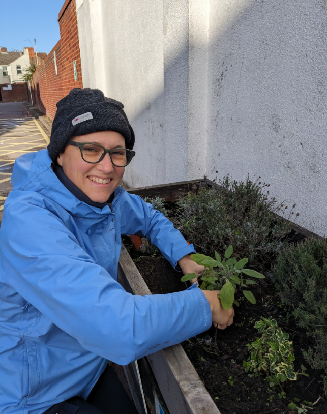 Volunteer form Fawcett Road Greening group with glasses, a black beanie, and a blue jacket planting or tending to plants in a small outdoor garden bed next to a white wall with a brick building in the background.