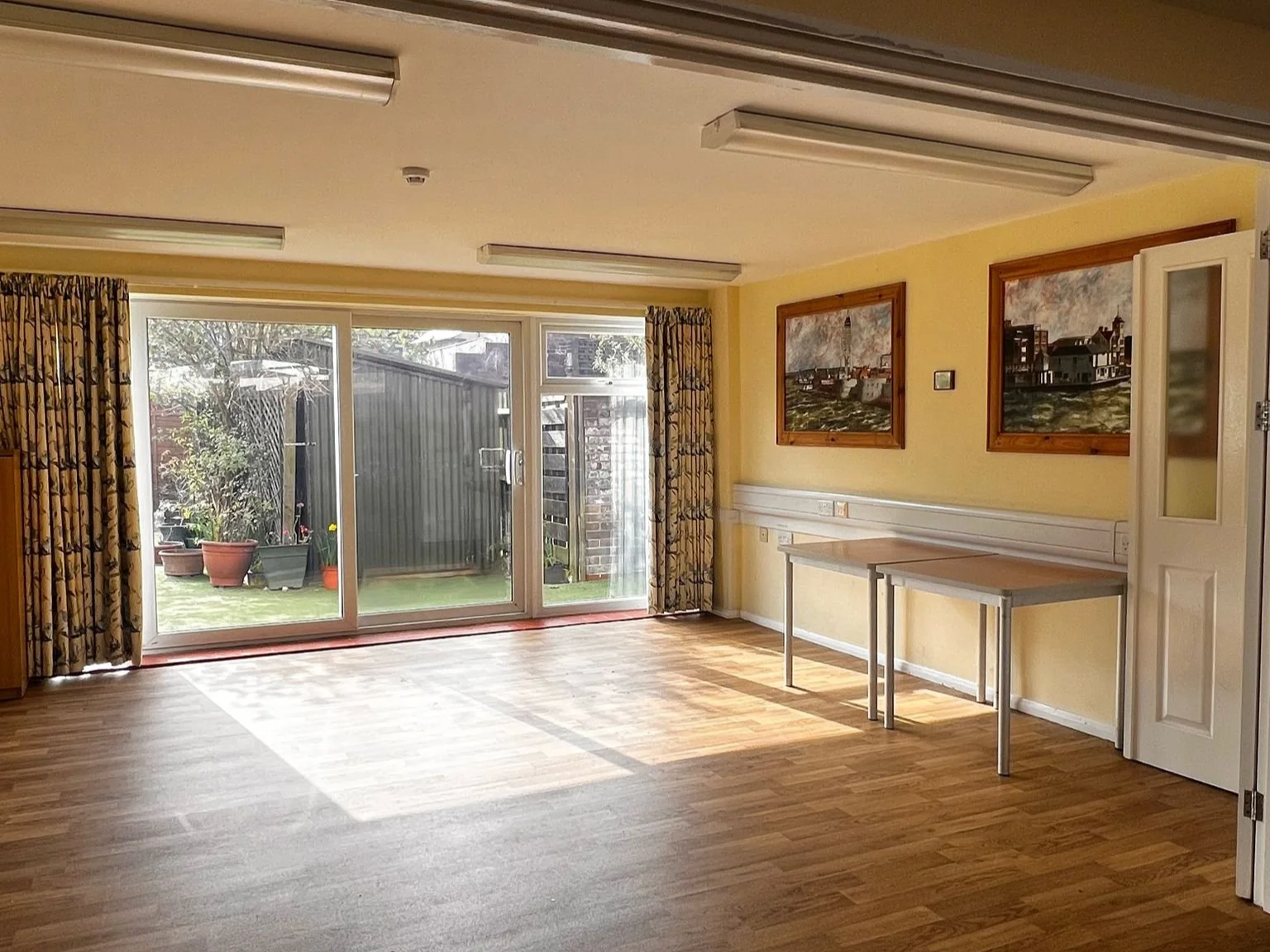 An empty room with wooden flooring, yellow walls, two framed paintings of a lighthouse and buildings, large glass sliding doors opening to a backyard with potted plants, and a partially open door on the right.