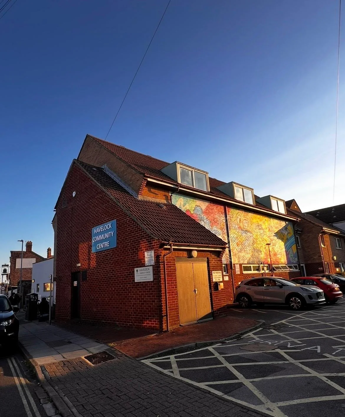 Brick building with signs indicating it is the Havelock Community Centre, parked cars, and a mural on the upper part of the wall, under a clear blue sky.