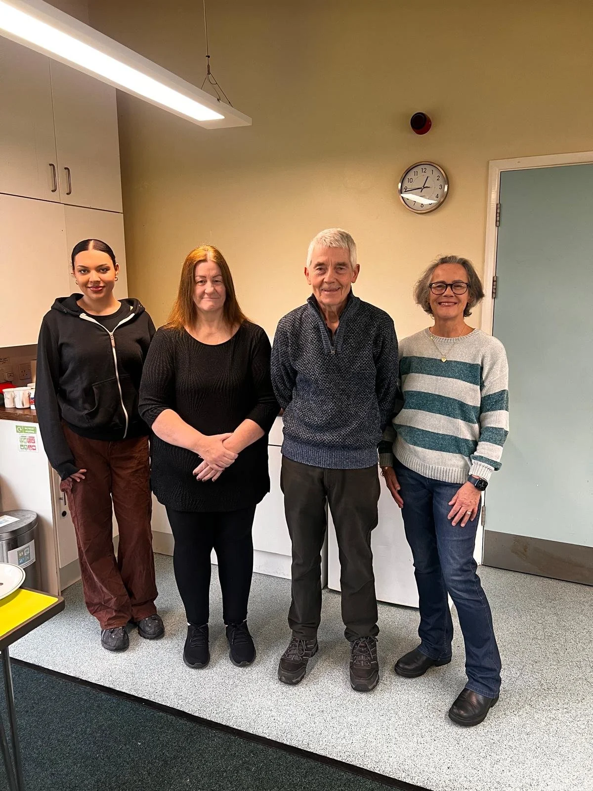 Group of four people standing in a room with kitchen cabinets and a clock on the wall.