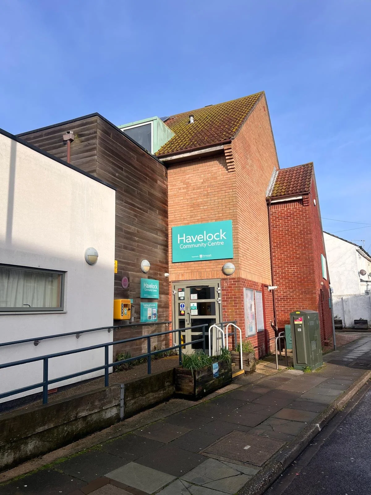 Exterior view of Havelock Community Centre with a blue sign, a white building, and a brick structure under a partly cloudy sky.