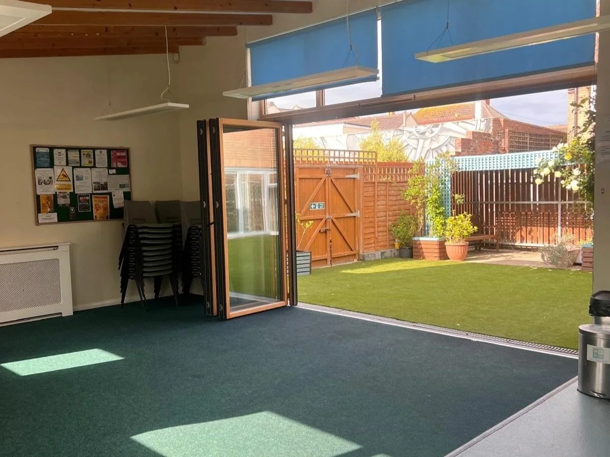 Interior room with green carpet, open door leading to a backyard with grass, potted plants, and a wooden fence, bulletin board on wall, chairs stacked in the corner, and natural light coming through the windows.