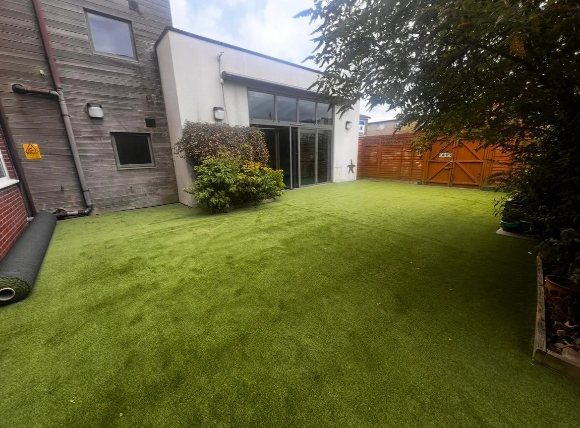 Backyard with artificial grass, bushes, a wooden fence, and a modern building with large windows and a sliding glass door.