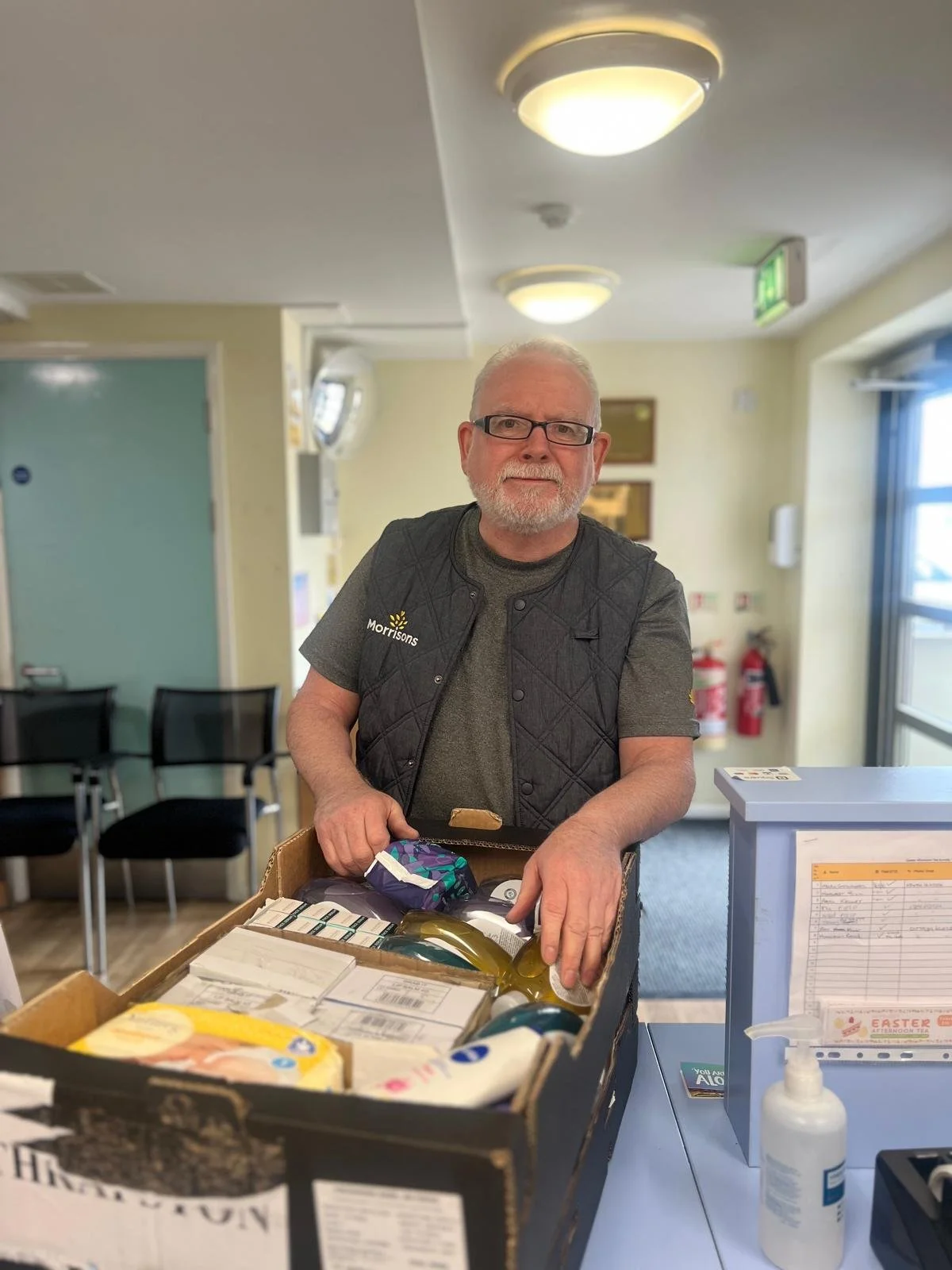 An man with glasses and a beard, wearing a gry shirt and a black vest that says 'Morrisons,' standing behind a counter with a large box of groceries in a store or cafe. The background shows chairs, a door, and a wall with fire extinguishers and fire alarm equipment.