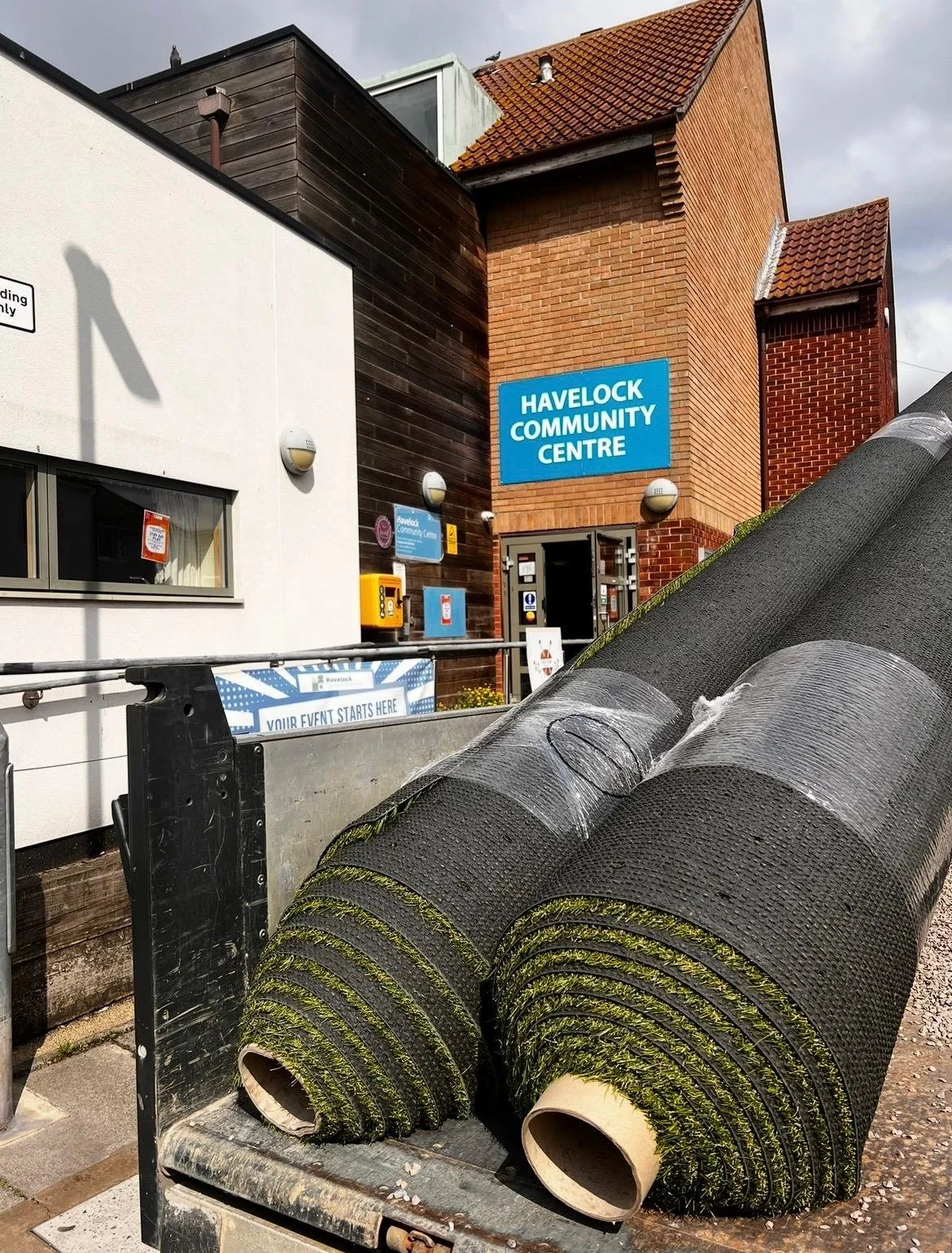 Construction rollers with moss on them in front of the Havelock Community Centre building with a blue sign, brick and wooden exterior, and cloudy sky.