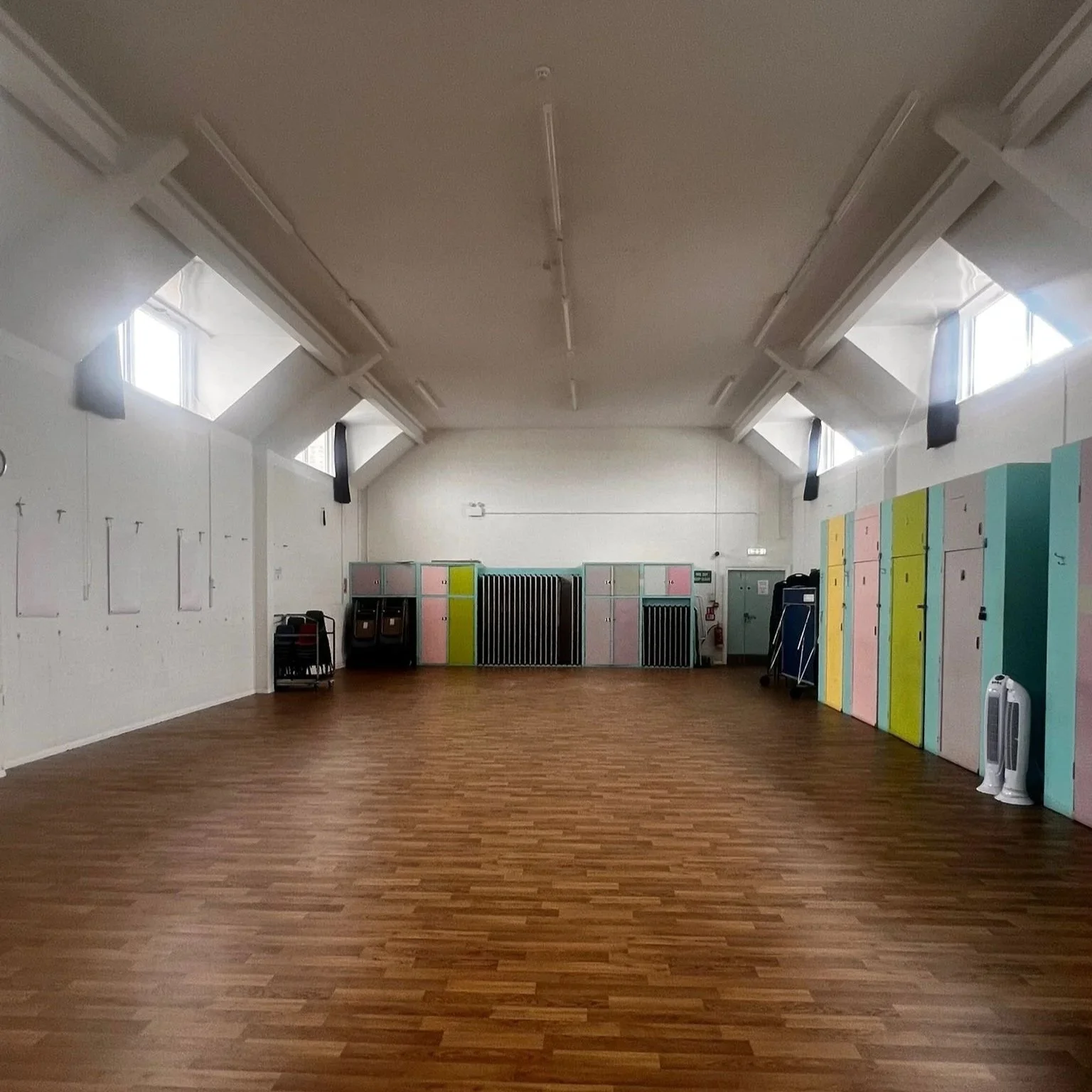 Empty room with wooden flooring, pastel-colored lockers on the right, and some black carts and a fan on the left side.