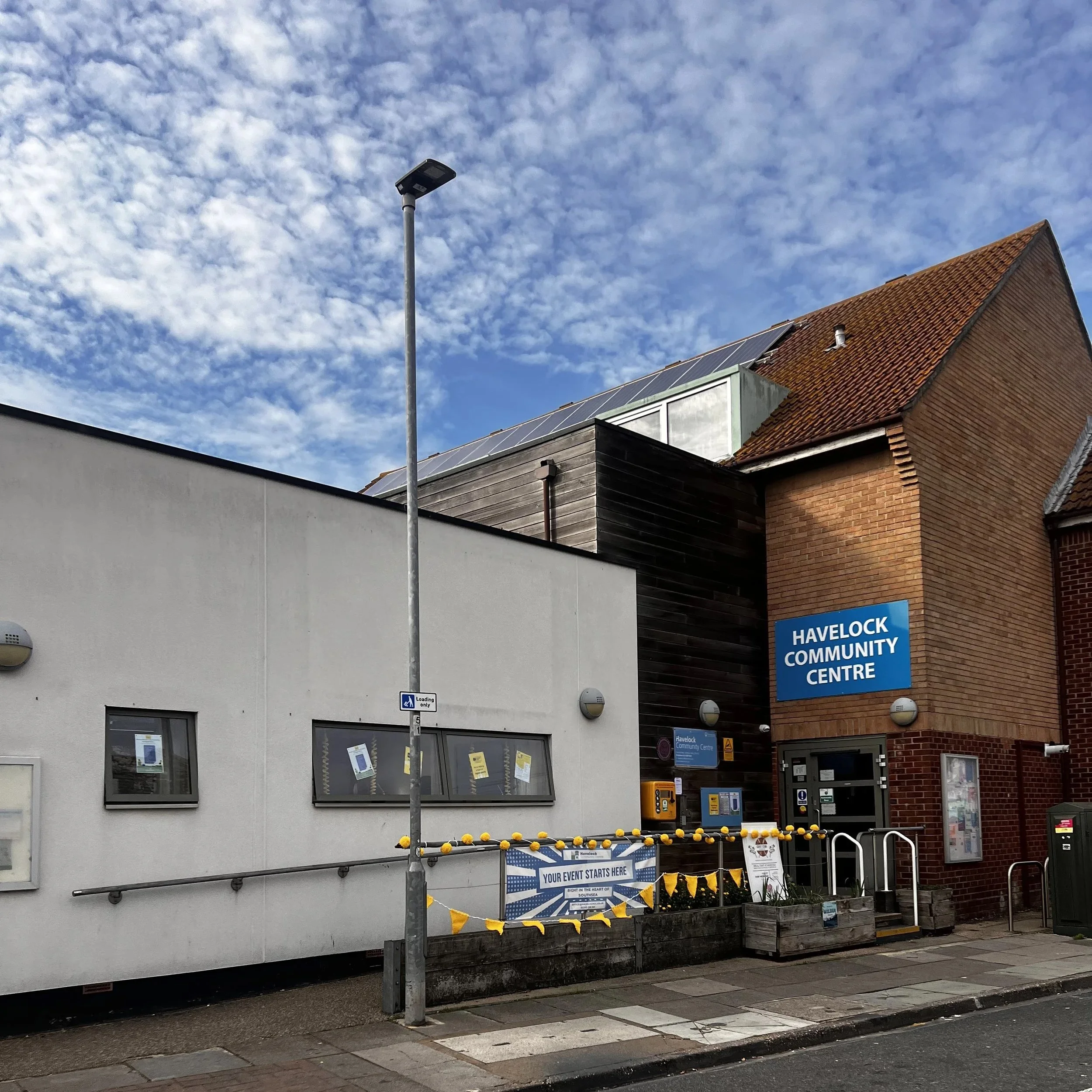 Exterior view of Havelock Community Centre with a blue sign, a white building, and a brick structure under a partly cloudy sky.
