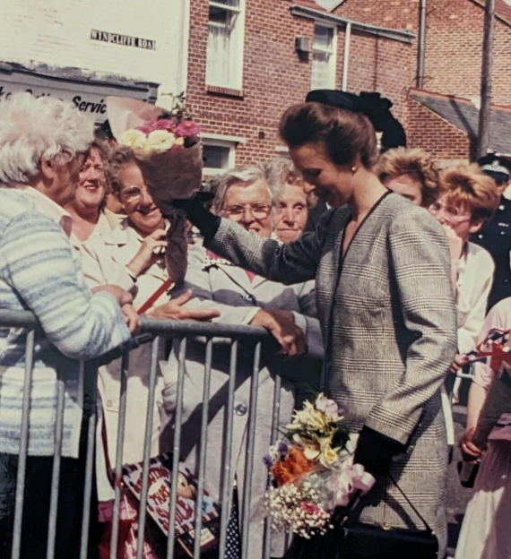 A young woman with a flower bouquet shaking hands with an elderly woman behind a metal barrier, surrounded by smiling onlookers on a street during a public event.