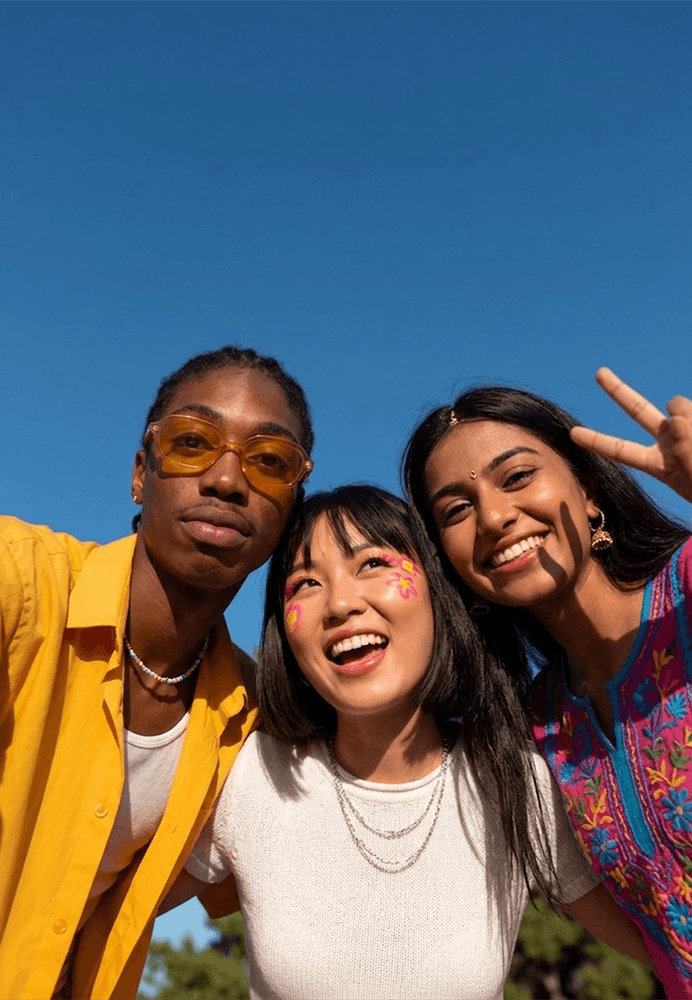 Three people leaning together beneath a blue sky, one wearing yellow sunglasses and another raising a peace sign.