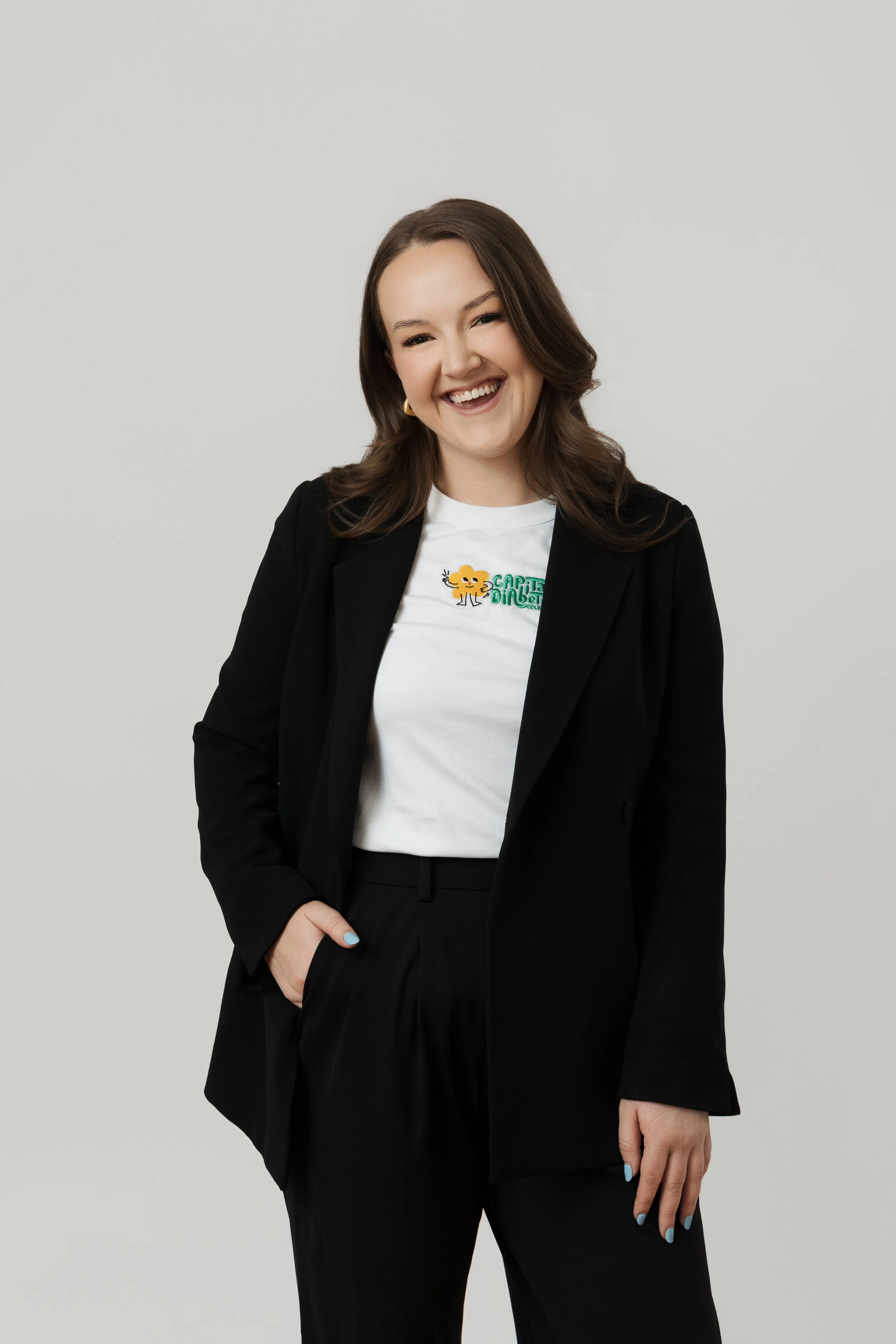 A smiling woman with long brown hair wearing a black blazer over a white t-shirt and black pants, with hand in pocket, standing against a plain light gray background.