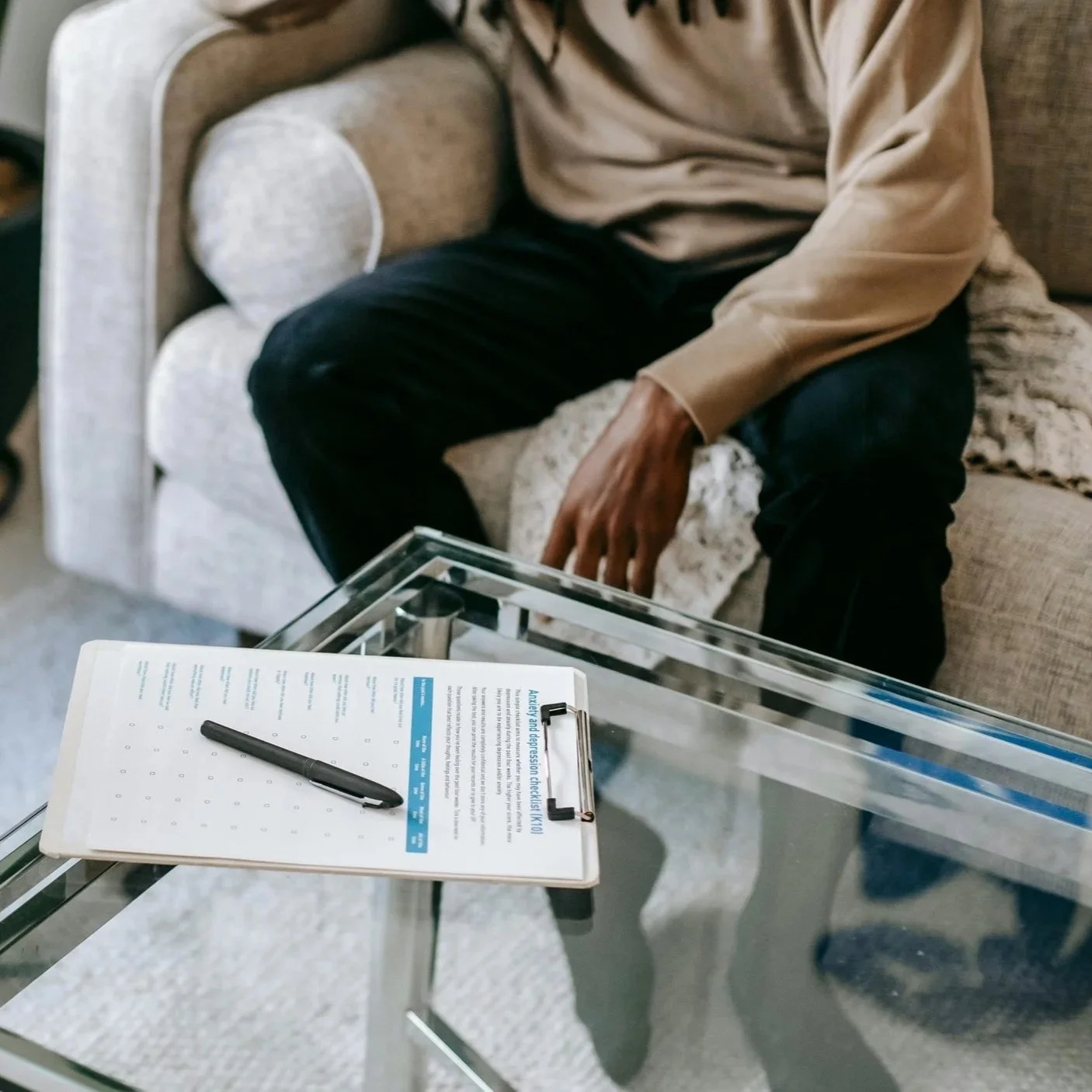 A person sitting on a beige sofa with their hand resting on a cushion. In front of them is a glass table with a clipboard, paper, and a black pen on it.