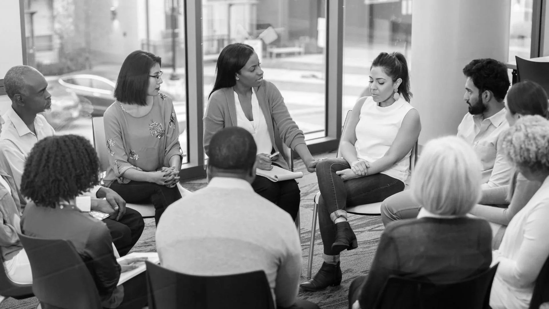 A diverse group of people in a meeting room, sitting in a circle, engaged in a discussion with a facilitator taking notes.