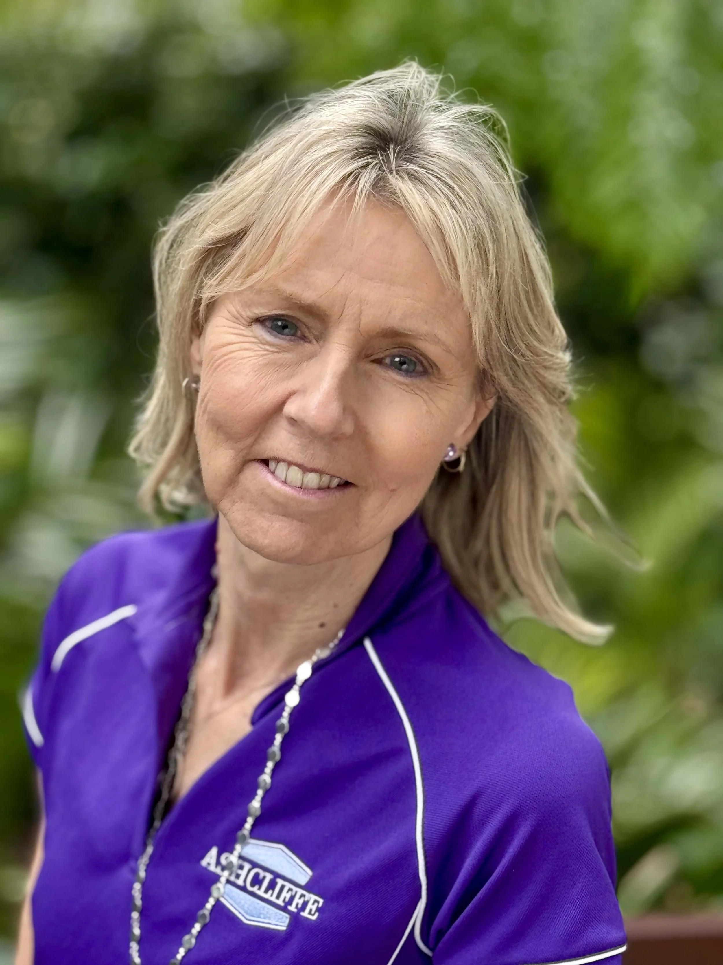 A woman with short blonde hair and blue eyes smiling, wearing a purple sports shirt with white piping and a necklace, outdoors with blurred green foliage in the background.